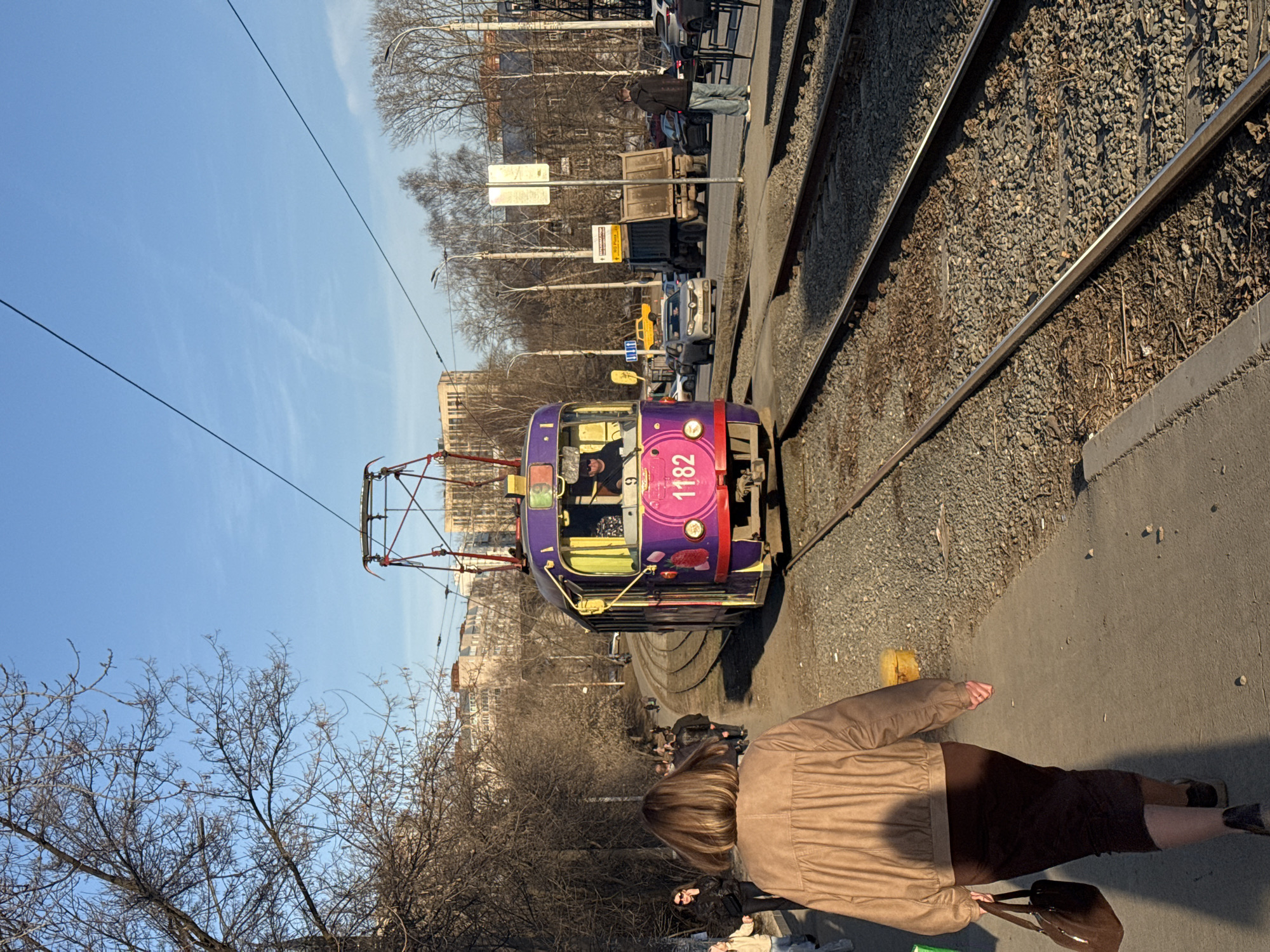 A purple and red tram, numbered 1182, is traveling along a track. A woman in a tan jacket and brown skirt is walking along the sidewalk. Other pedestrians are visible in the background. The scene is set in Izhevsk, Russia. Tracks of the tram run along gravel. Bare trees can be seen in the background. Cars are on the road next to the tracks. There are street lamps along the road. The sky is blue.