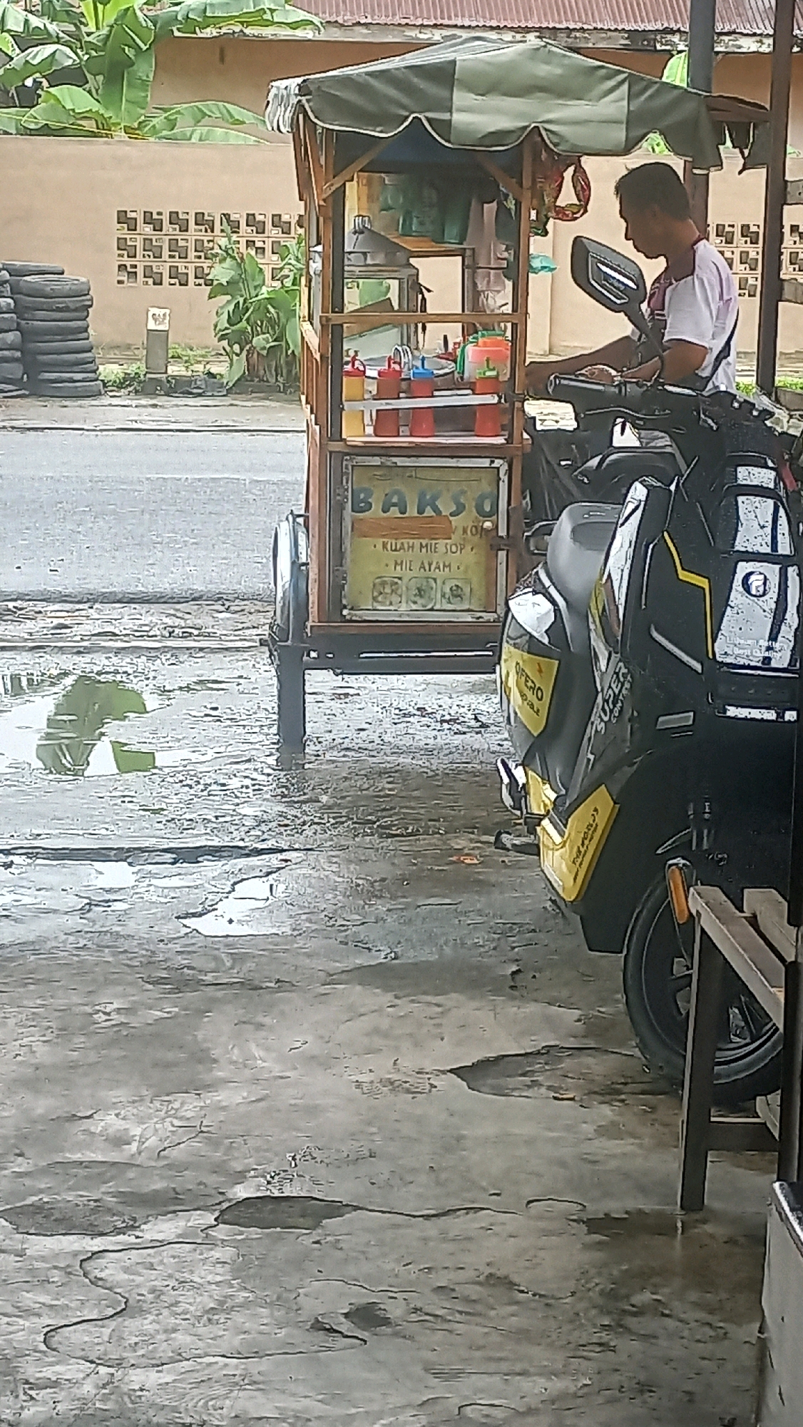 A man, wearing a white t-shirt with a maroon collar and sleeve trim, is sitting on a black scooter. The scooter is parked next to a food cart that sells "Bakso" (Indonesian meatball soup). The cart has a wooden frame and a green awning. Condiments and utensils are visible on the cart. The ground is wet and reflects the surroundings, suggesting it has recently rained. In the background, there is a wall with decorative concrete blocks, lush green plants, and a stack of tires. The scene appears to be in a street or roadside location in the city of Medan, Indonesia.