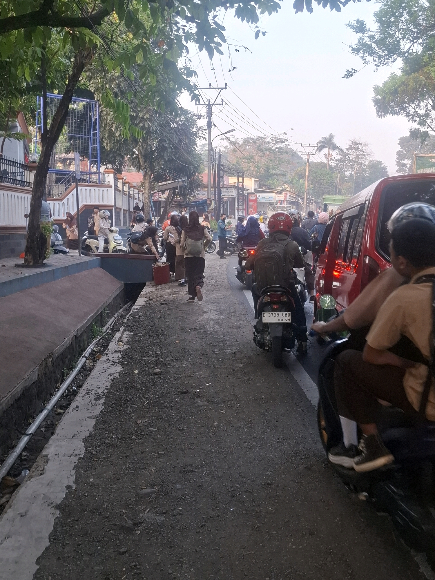 The image depicts a street scene in Sirnagalih, Indonesia, with a focus on a busy road populated by pedestrians and motorcyclists. Several students, identifiable by their school uniforms and backpacks, are walking along the side of the road and riding motorcycles. One student is seen near a red trash bin, possibly disposing of waste. A red vehicle, possibly a van, is stopped on the right side of the road, with several motorcyclists behind it. Trees line the street, and utility poles with numerous wires are visible in the background, suggesting an urban or semi-urban environment. The road surface is gravelly, and a concrete drainage ditch runs along the left side of the image. The overall atmosphere appears to be that of a typical day in a residential or school area.