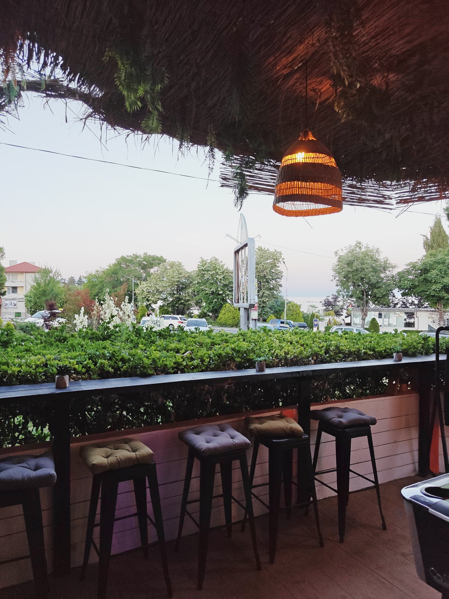 This is an outdoor photograph of a bar or restaurant area in Tsarevo, Bulgaria. The perspective is from a seated position looking out. In the foreground, a dark wooden counter with four bar stools is visible. The stools have cushioned seats in various colors, including grey, yellow, and dark grey. Behind the counter, a dense green hedge forms a barrier. Beyond the hedge, a street scene unfolds with trees, buildings, and parked cars. The sky is overcast and pale, suggesting late afternoon or early morning. A decorative woven lampshade hangs from the thatched roof overhead, casting a warm glow. The overall impression is of a relaxed, casual dining or drinking establishment.