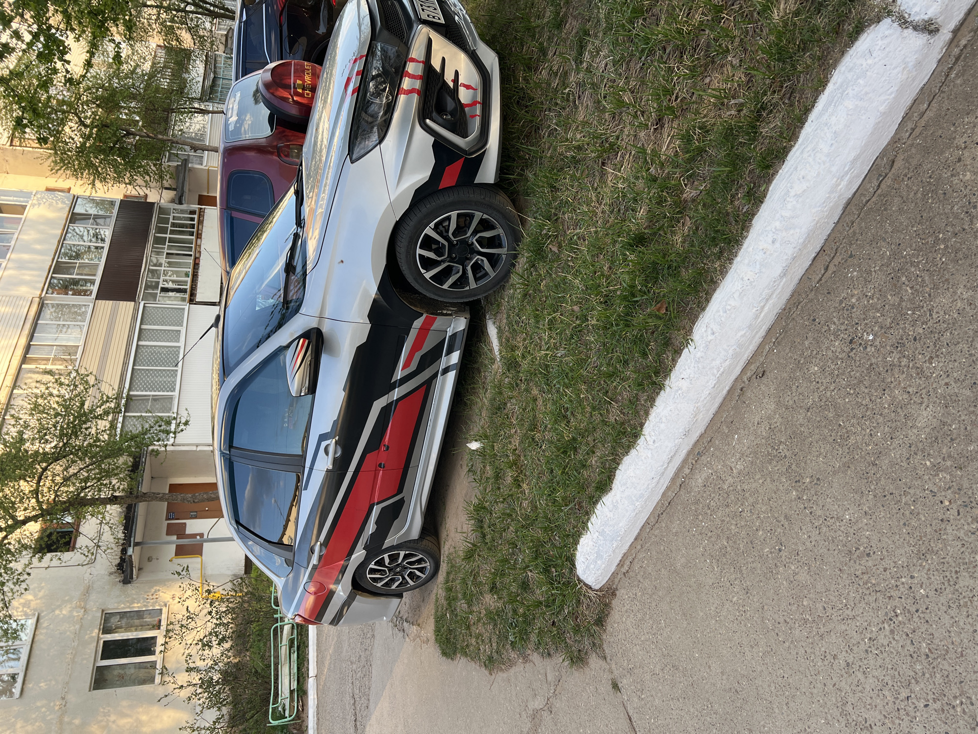 The image shows a silver car with red and black racing stripes parked on the edge of a grassy area in Noviy, Russia. The car is next to a concrete sidewalk with a painted white curb. In the background is a multi-story apartment building with windows and balconies. Other cars are parked behind the silver car. The photo appears to have been taken during daylight hours.