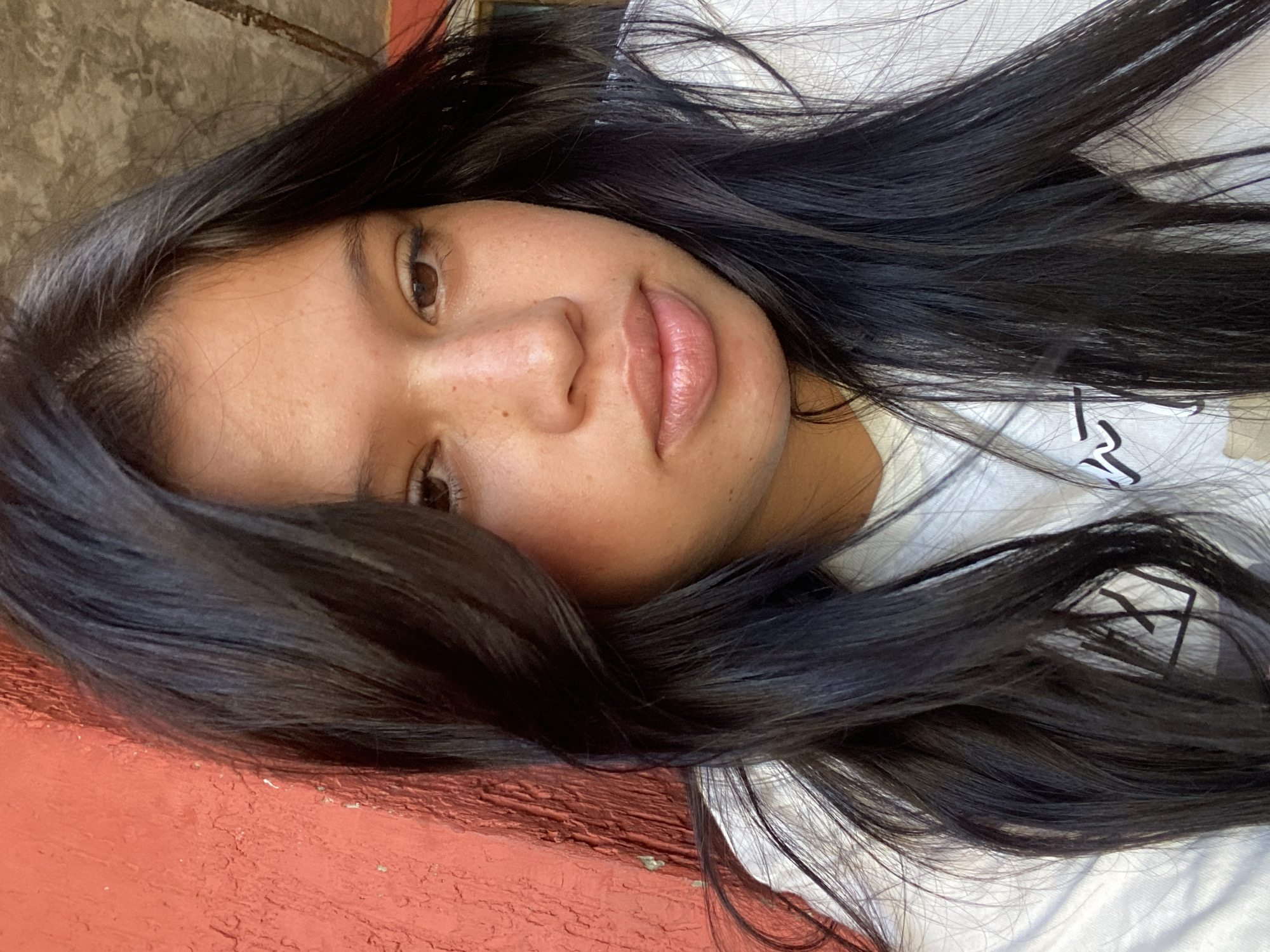 The image is a close-up portrait of a woman lying down. She has dark, long hair that surrounds her face. Her skin is light brown, and she has brown eyes. She is wearing a white shirt. The background consists of a red surface and a concrete structure. The location in the city of Juan Griego, Venezuela is not discernible from the image.