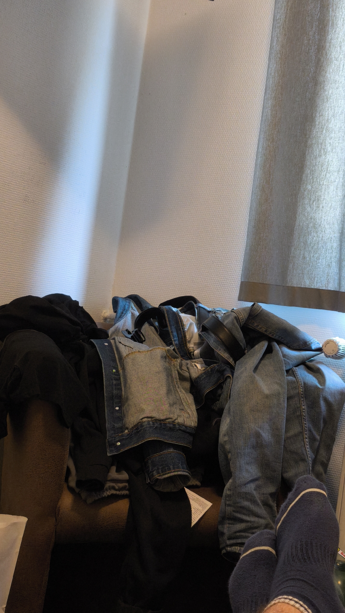 The image shows a pile of clothes on a brown upholstered surface, likely a couch or chair. The clothing includes a black garment, a blue denim jacket, and blue denim jeans. A pair of feet wearing dark blue socks with white stripes is visible in the foreground. A white piece of paper is partially visible amidst the clothing. The background includes a white wall and a light-colored curtain. The location is Unknown, Unknown.