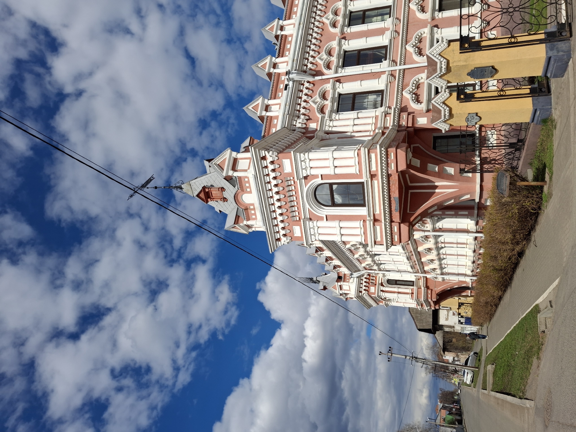 The image showcases a building located in Sarapul, Russia. The architectural style is characterized by red and white detailing. The building features multiple windows and decorative elements. Overhead power lines are visible against a blue sky with clouds. A sidewalk and street flank the building. A portion of other buildings and trees can be seen in the background.
