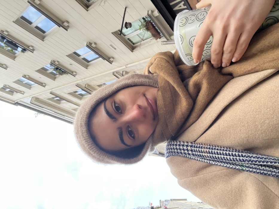 An individual, identified as female, is depicted in a close-up selfie taken outdoors in an urban setting. She has dark hair, dark eyebrows, and is looking directly at the camera with a slight smile. She wears a light brown knitted hood, a camel-colored scarf wrapped around her neck, and a camel-colored coat. A black and white houndstooth patterned strap, likely from a bag, is visible over her left shoulder. In her right hand, she holds a clear plastic cup containing a light green beverage, fitted with a lid and straw. The cup is decorated with light green smiley face patterns.
The background features a multi-story building constructed from light-colored stone or stucco, typical of Haussmannian architecture found in Paris, France. The building has numerous rectangular windows with light-colored frames. Some windows are adorned with dark-colored planter boxes containing green plants. A dark, ornate hanging lantern fixture is visible on the building's facade to the lower right. A partial black sign with white lettering, showing "AL", is discernible behind the cup. The sky above is uniformly light gray or white, indicating an overcast condition. The scene is consistent with a street or pedestrian area within Paris, France.