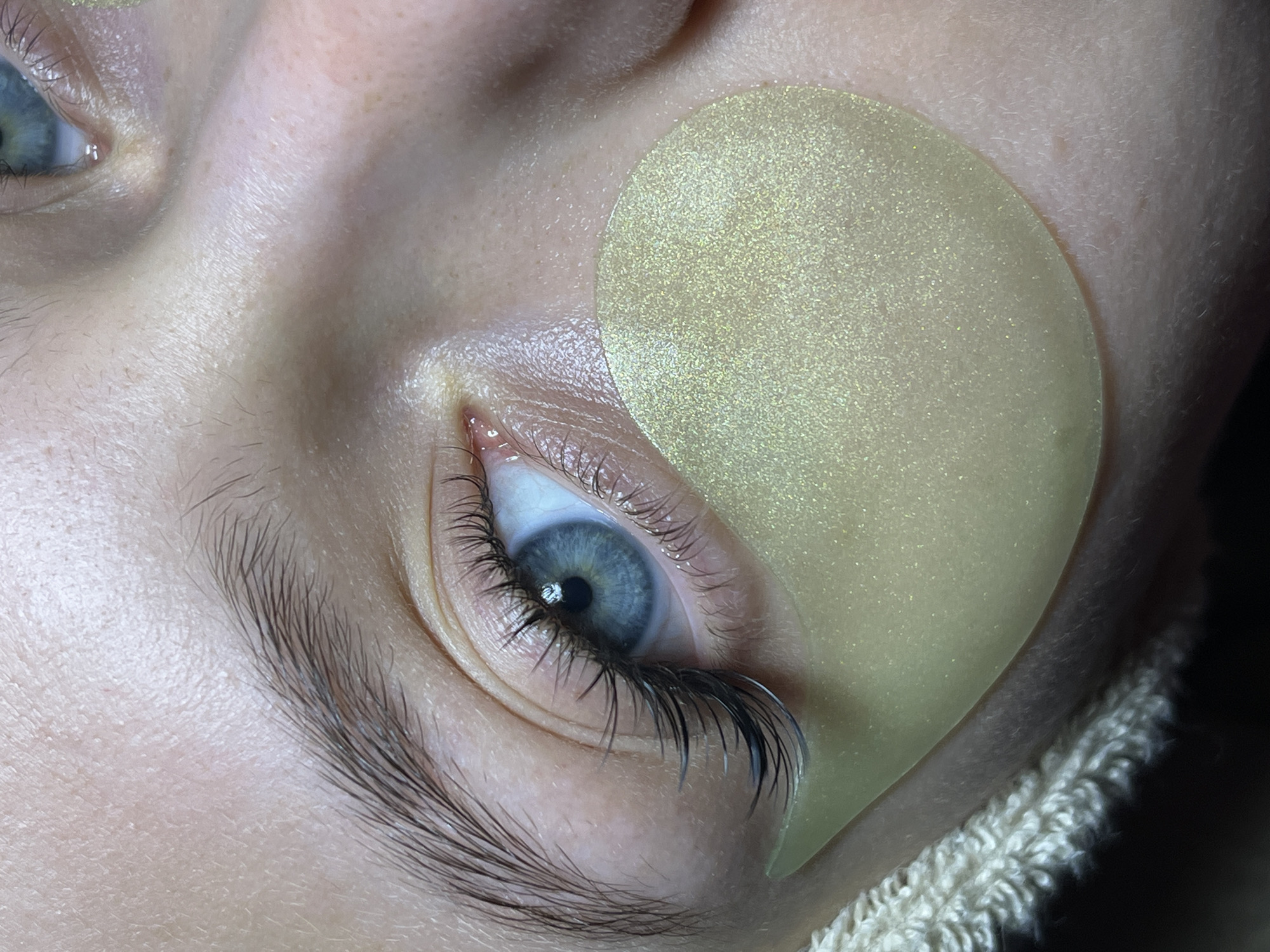 A close-up image shows a person's eye with an under-eye mask applied. The eye is open and blue. The eyelashes appear to be recently extended. The mask is a light yellow color and covers the area under the eye. The person's eyebrow is visible and has dark hair. No specific details indicate the location as Фоки, Russia. The background is generally blurred, but some fabric can be discerned under the lower face.