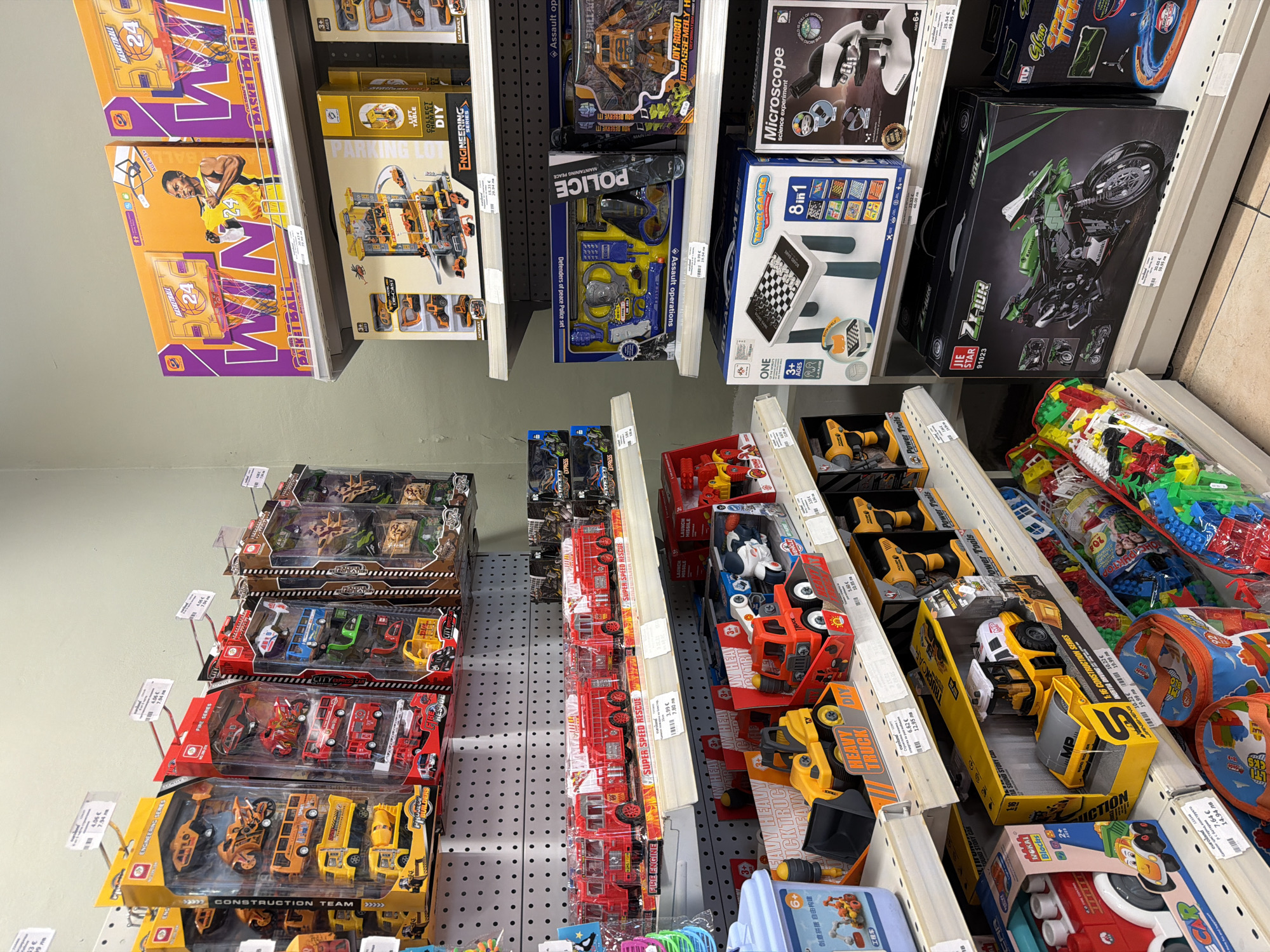 This is an indoor, eye-level, medium shot of shelves stocked with toys in a store. The shelves are white, metal, and have price tags and labels on some of the toys. The store appears to be well-lit and organized. The toys are various types of vehicles, including construction vehicles, fire trucks, and motorcycles, as well as some educational toys like a microscope and a chess set. The overall impression is a typical toy store environment. The image was taken in Pazardzhik, Bulgaria.