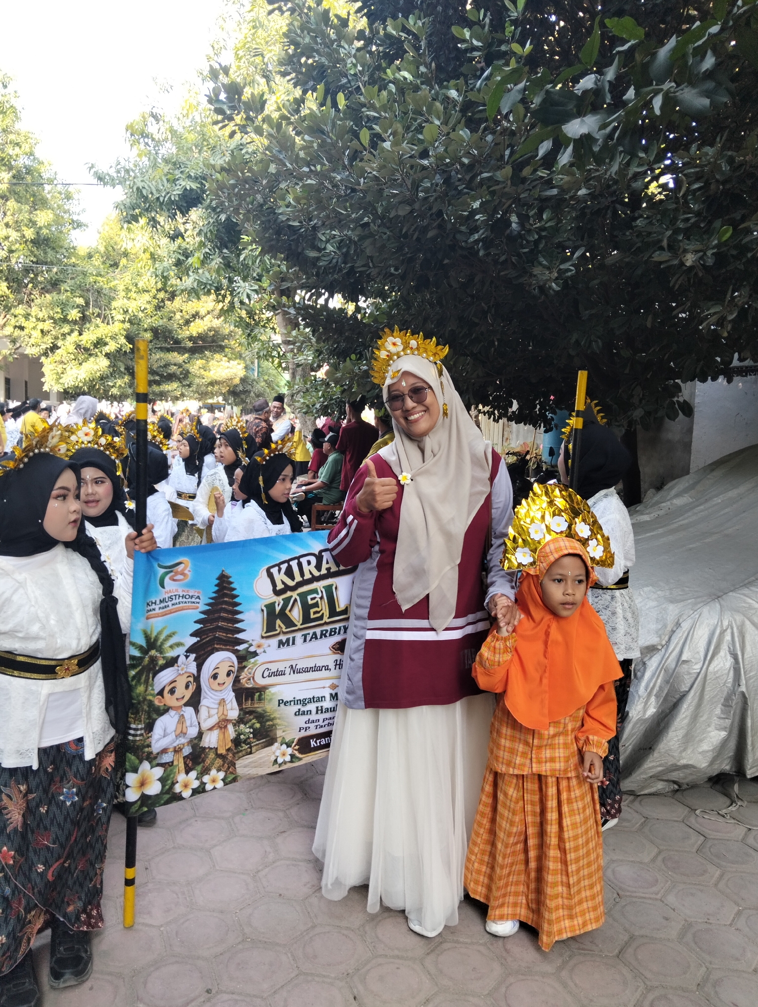 In Kranji, Indonesia, a group of children dressed in traditional attire, including headscarves and ornate gold headdresses adorned with white flowers, participate in a parade. They stand with a banner that features cartoon depictions of a boy and girl in similar outfits, a Balinese temple, and palm trees. The banner also contains text in Indonesian that mentions "Peringatan Maulid Nabi" and "MI Tarbiyah." An adult woman, wearing a headscarf and sunglasses, stands among the children, holding the banner and giving a thumbs-up. The ground is paved with hexagonal tiles. The background consists of trees and buildings, suggesting an outdoor urban setting.