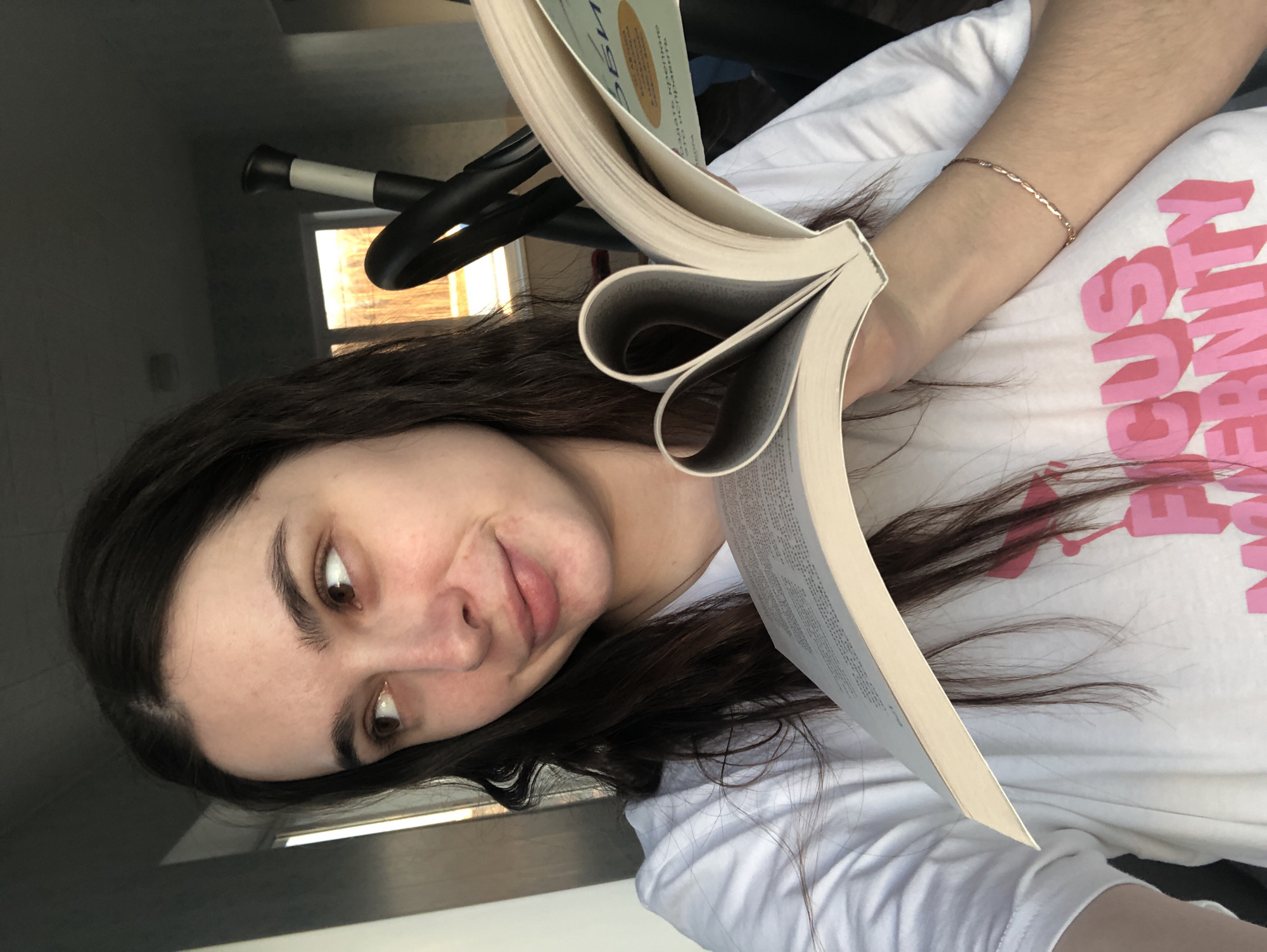 A young woman with dark hair and light skin holds an open book. The book pages are folded into heart shapes. She wears a white t-shirt with pink lettering. A gold bracelet is on her left wrist. A black and white elliptical machine or similar exercise equipment is visible in the background, near a window. The image appears to be taken indoors. There is no evidence to indicate this is in the city of Noviy, Russia.