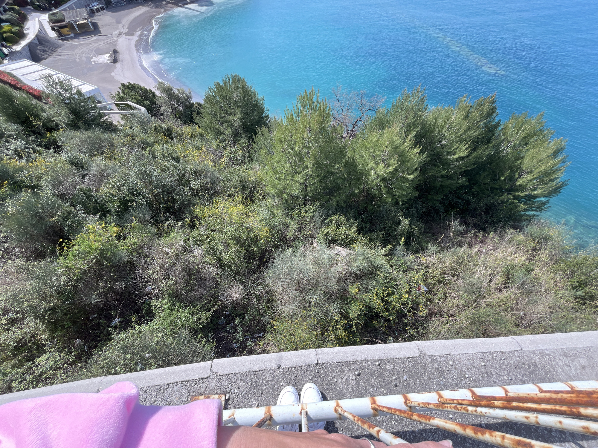 A high-angle photograph captures a steep, vegetated cliffside overlooking a beach and the Adriatic Sea in Budva, Montenegro. In the immediate foreground, the feet of an individual, wearing white sneakers, are visible positioned directly behind a white metal railing heavily marked with reddish-brown rust. A pink, fuzzy textile object rests on a concrete ledge to the left. The midground consists of a dense, descending slope covered in various green shrubs and bushes, with pockets of yellow flowering plants. Scattered pieces of litter, including plastic bottles and bags, are visible amongst the vegetation. In the background to the left, a dark sand and pebble beach is visible, with several modern white-roofed structures and a winding paved path. The expanse of the Adriatic Sea dominates the background, displaying clear turquoise water near the shore and deeper blue further out, with faint submerged formations visible. Numerous green trees, resembling pines, grow along the lower parts of the cliff leading to the sea.