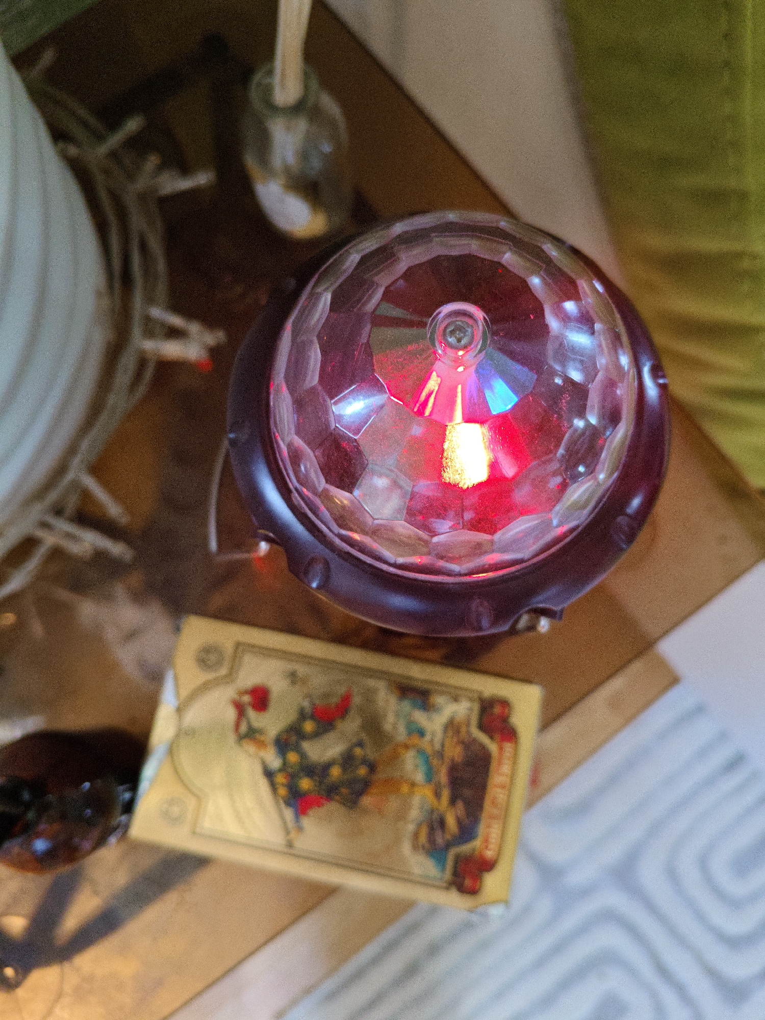 A close-up, high-angle shot shows a multifaceted disco ball light emitting red and blue hues, resting on a wooden surface. Beneath the disco ball sits a deck of tarot cards, featuring golden borders and colorful illustrations of figures. To the left, a white coiled object, possibly a rope or string of lights, is visible, along with a dark, glossy object with blue speckles. In the background, a small glass bottle with a wooden stick is partially seen, and a soft, textured surface, possibly upholstery, is present. The lighting in the scene is dim, with the disco ball being the primary light source. The image is not identifiable as being located in Rostov-on-Don, Russia.