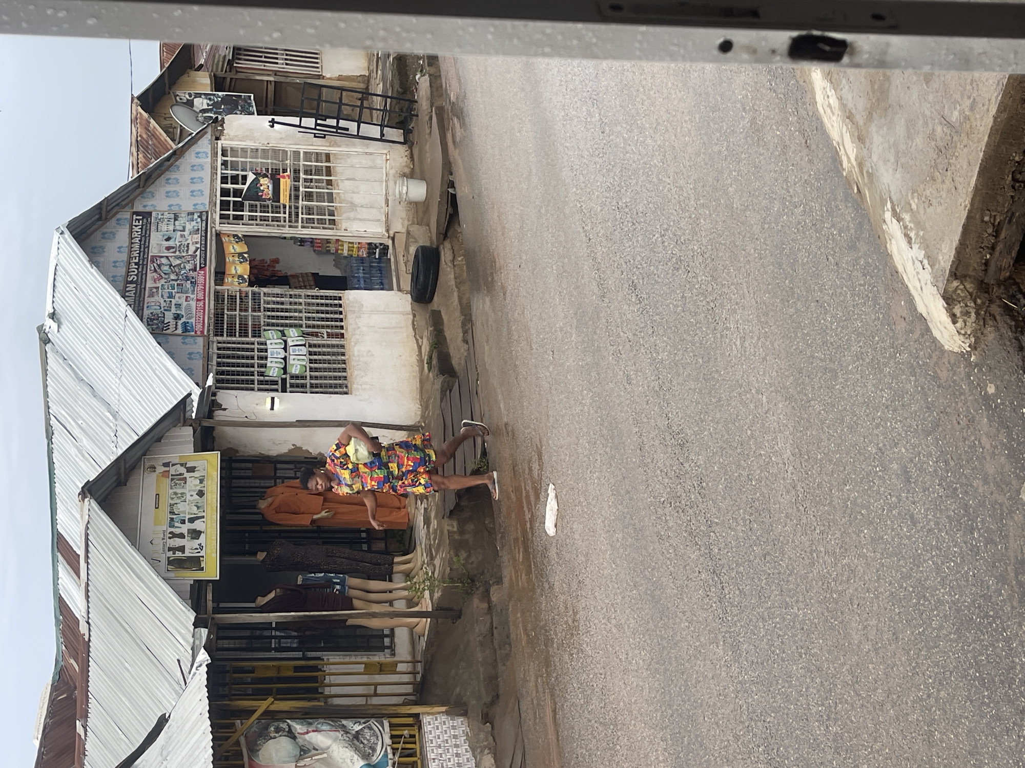 The image captures a street scene in a commercial or market area, likely in Abuja, Nigeria, under an overcast sky during the daytime. The view is from a slightly elevated perspective, looking down at a paved road bordered by buildings housing various shops.
In the mid-ground, a person wearing a vibrant, multi-colored short-sleeved shirt and shorts is standing on the sidewalk, near the storefronts. Beside them, several mannequins are displayed, dressed in different clothing items, indicating a clothing retail business.
The buildings lining the street feature corrugated metal roofs and windows secured with grilles or bars. One prominent building has a large sign that clearly reads "AMAN SUPERMARKET," adorned with images of products and including phone numbers "09032203159" and "09079159914." Another sign, appearing to be a poster or framed display, shows multiple smaller images, possibly of clothing styles, with some unreadable text below. A satellite dish is visible on the upper part of one of the buildings.
On the ground next to the road, a black tire and a white bucket are present. The road itself is gray asphalt. The scene depicts a typical local retail environment, with no discernible specific activity beyond the general presence of people and shops.