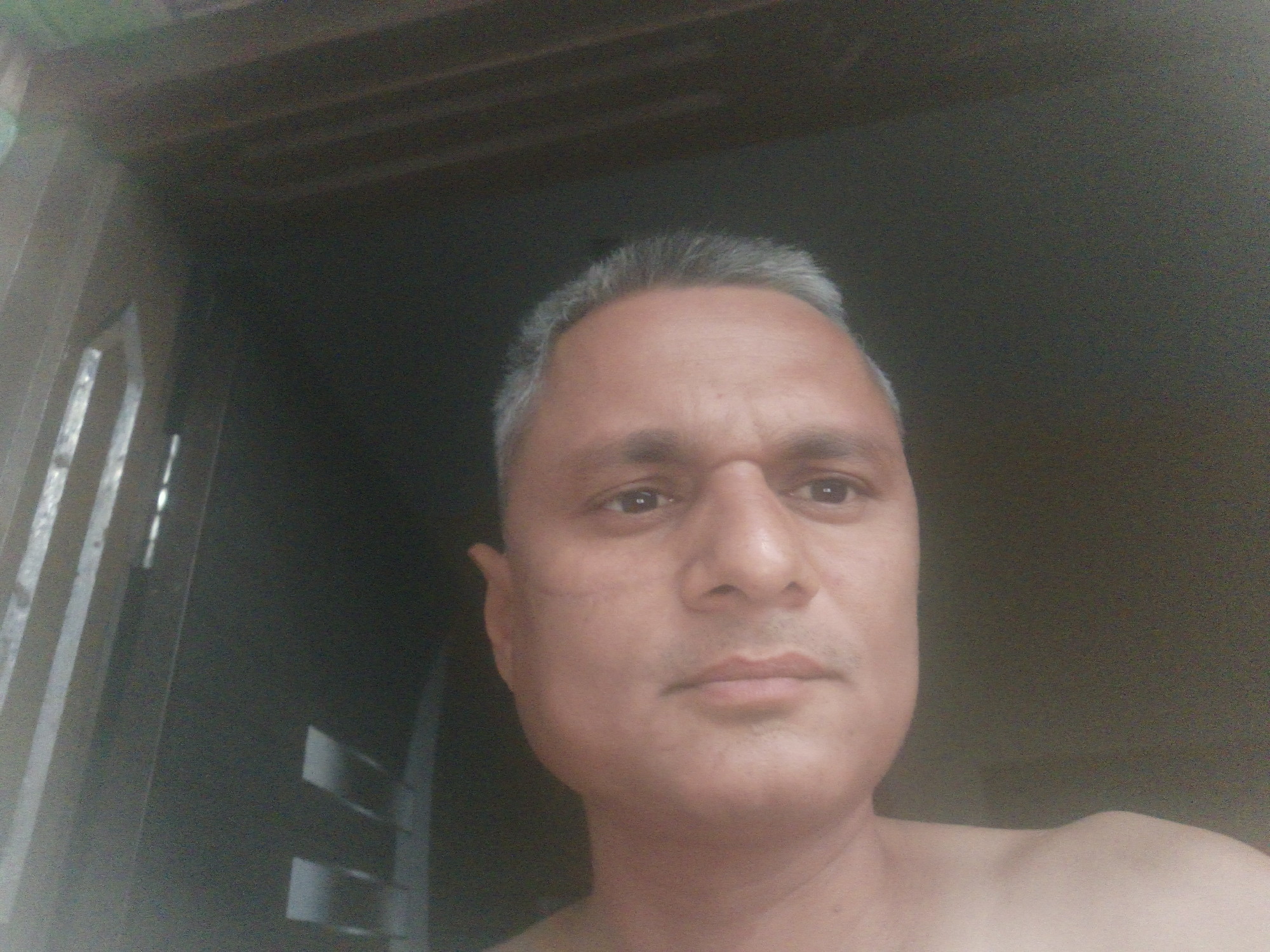 A close-up, low-angle self-portrait of a middle-aged man with short, graying hair and light brown skin. He has a serious expression. He appears to be indoors, standing in front of a dark doorway or opening. No specific details visible indicate the location is Dhandhuka, India.