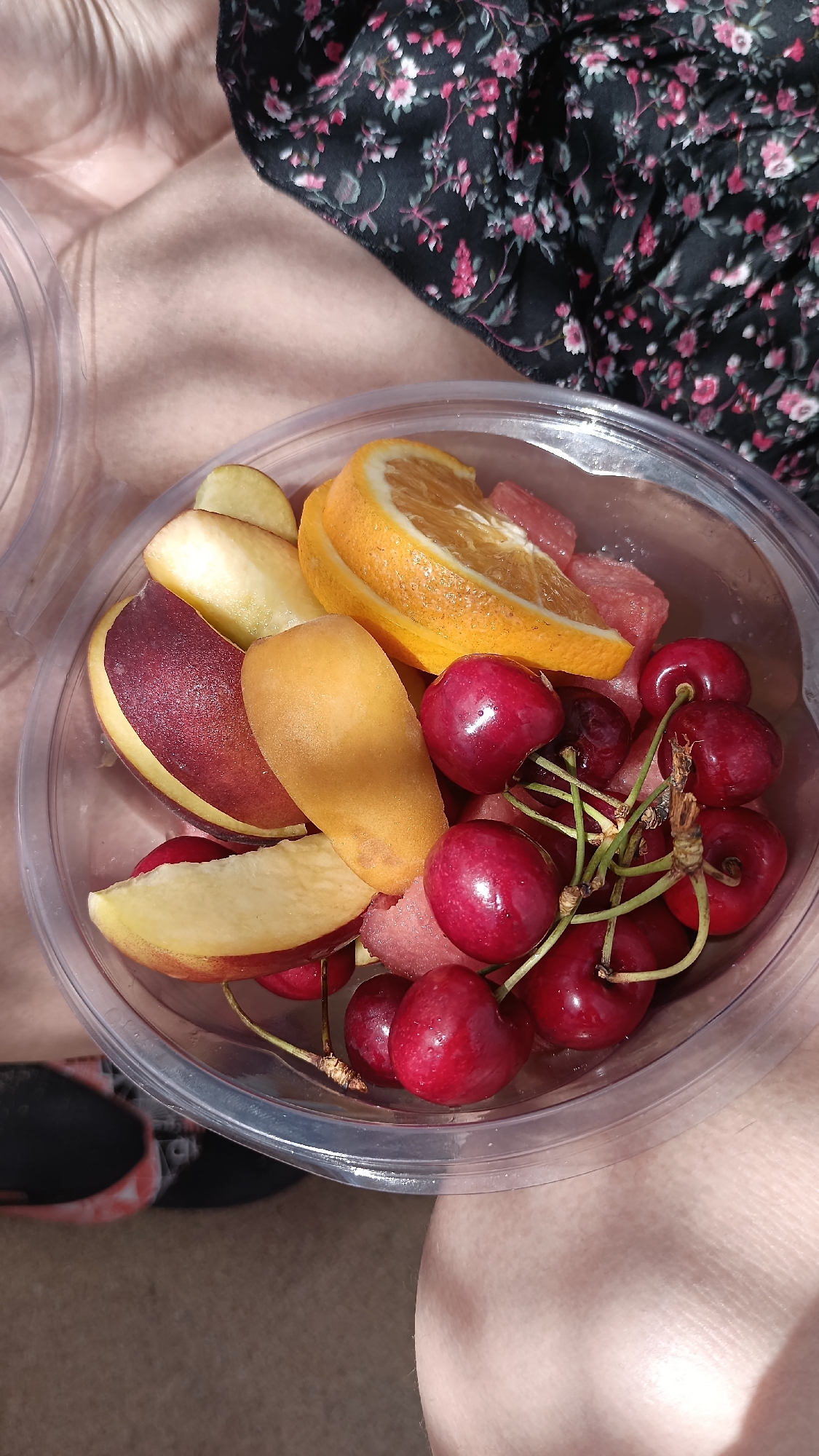 A close-up, overhead shot shows a clear plastic container filled with a colorful assortment of fresh fruit. The container rests on a person's lap, indicated by the visible legs and the edge of floral fabric, possibly a skirt or dress, in the background. The fruit selection includes bright red cherries, some still attached by their stems, slices of juicy orange, pieces of what appear to be apple or peach, and a few chunks of pink watermelon. The lighting suggests it is daytime, possibly a sunny afternoon, as evidenced by the strong shadows and highlights on the fruit and skin. The scene is casual and outdoors, with the texture of the ground visible beneath the person's legs. The overall impression is one of a refreshing, healthy snack enjoyed in a relaxed setting, likely in Nea Peramos, Greece, given the location context.