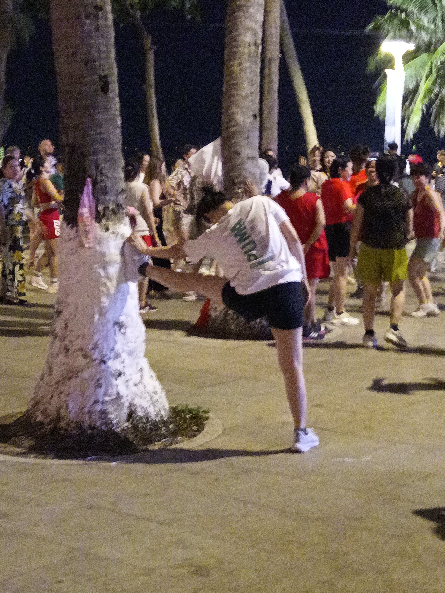 The image depicts a nighttime outdoor scene, likely in Nha Trang, Vietnam, given the presence of palm trees and the casual attire of the individuals. In the foreground, a woman wearing a white t-shirt with the word "PUMA" visible on the back and black shorts is performing a stretching exercise. She is facing away from the camera, with her right leg extended behind her and her left leg bent. To her left, a thick, textured tree trunk wrapped with a white, fabric-like material stands prominently. Behind the woman, a crowd of people are gathered, illuminated by artificial lighting. Many are wearing brightly colored clothing, predominantly red. The background includes more palm trees and a bright, modern lamppost. The ground appears to be paved, possibly a sidewalk or public area. The atmosphere suggests a social gathering or an organized event, possibly a fitness class or celebration, taking place along a promenade or beach area.