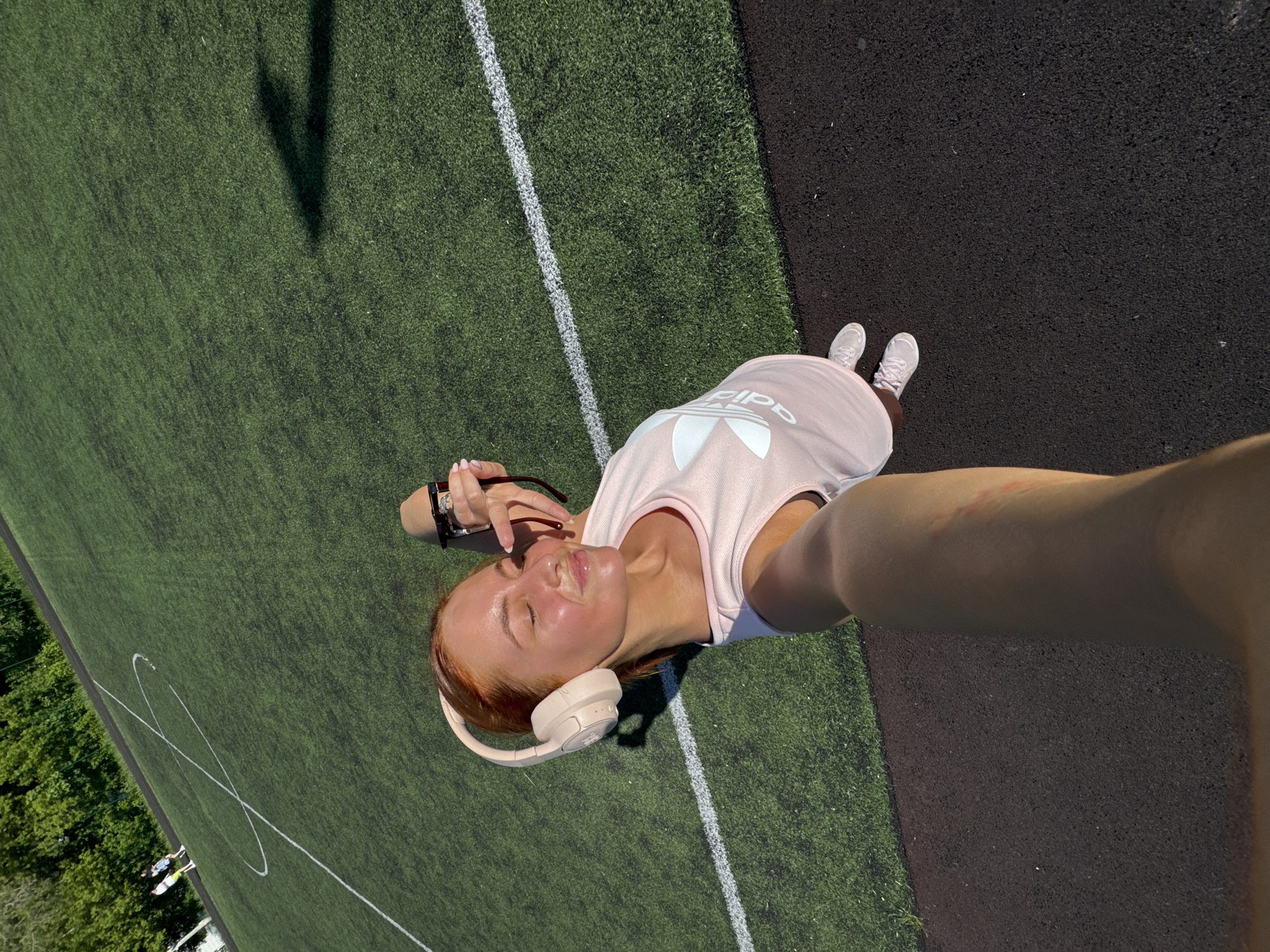 A woman stands on a black and green sports field, likely astroturf. She is wearing a pink Adidas t-shirt, white sneakers, and beige headphones. She holds sunglasses in her left hand and appears to be taking a selfie with her right arm extended. A white line marks the edge of the green field. Part of the image includes the shadow of a tall object on the field, as well as trees in the background. This photo may be taken at a sports complex or stadium in Chaikovsky, Russia.