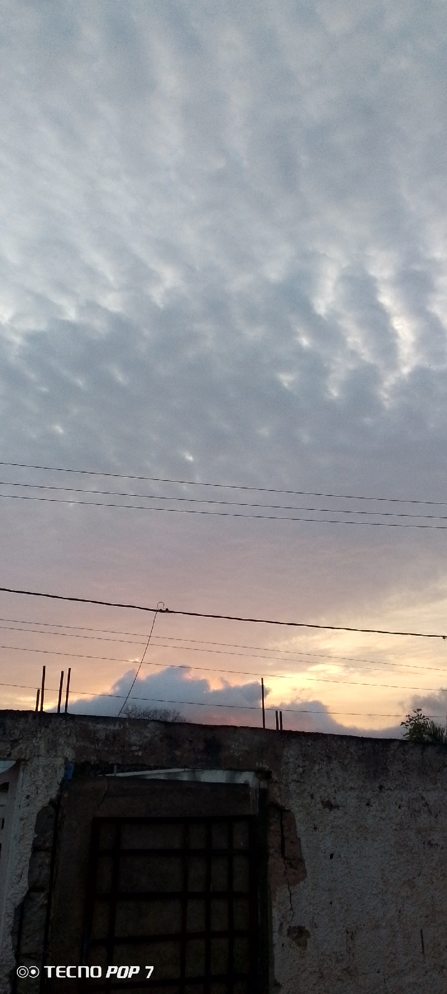 The photograph shows a sunset in Altagracia, Venezuela, taken from ground level, focusing on a dilapidated structure with a barred window. The upper portion of the frame is filled with a cloudy sky exhibiting hues of grey, white, orange, and pink, indicative of the setting sun. Several electrical wires traverse the sky horizontally. The foreground is dominated by a weathered wall with visible damage and a dark, barred window. The bottom left corner of the image displays the text "TECNO POP 7".