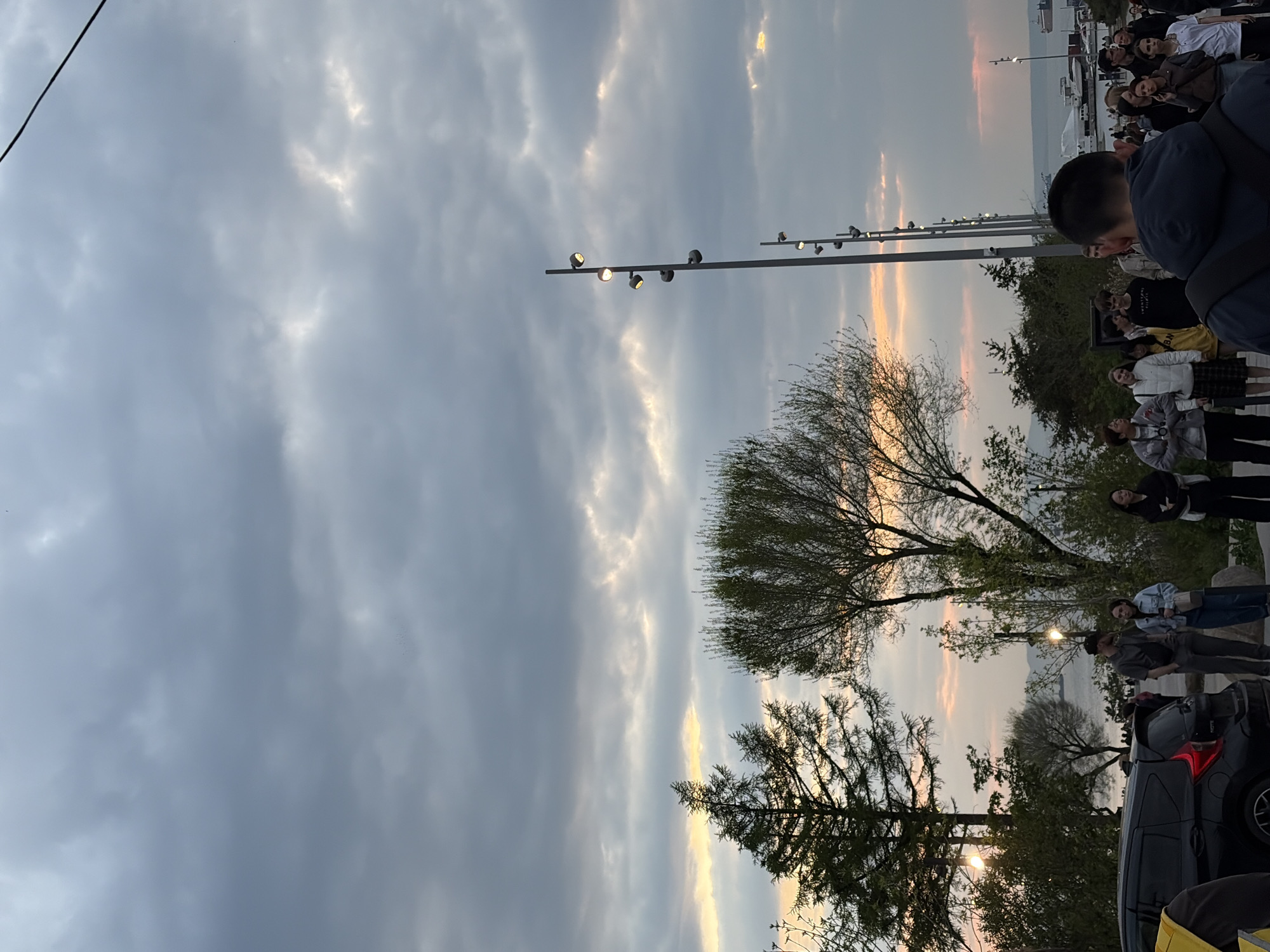 A medium outdoor shot, taken from a low angle, captures a cloudy sky in Vladivostok, Russia. The sky is filled with soft, grey clouds. Below, a line of modern streetlights, evenly spaced, adds artificial light to the scene. A row of trees with budding leaves and evergreens lines the horizon. A crowd of people are gathered in the background, their attention likely focused on something out of frame. Portions of cars are visible in the foreground. The scene is bathed in the subdued light of a cloudy evening.