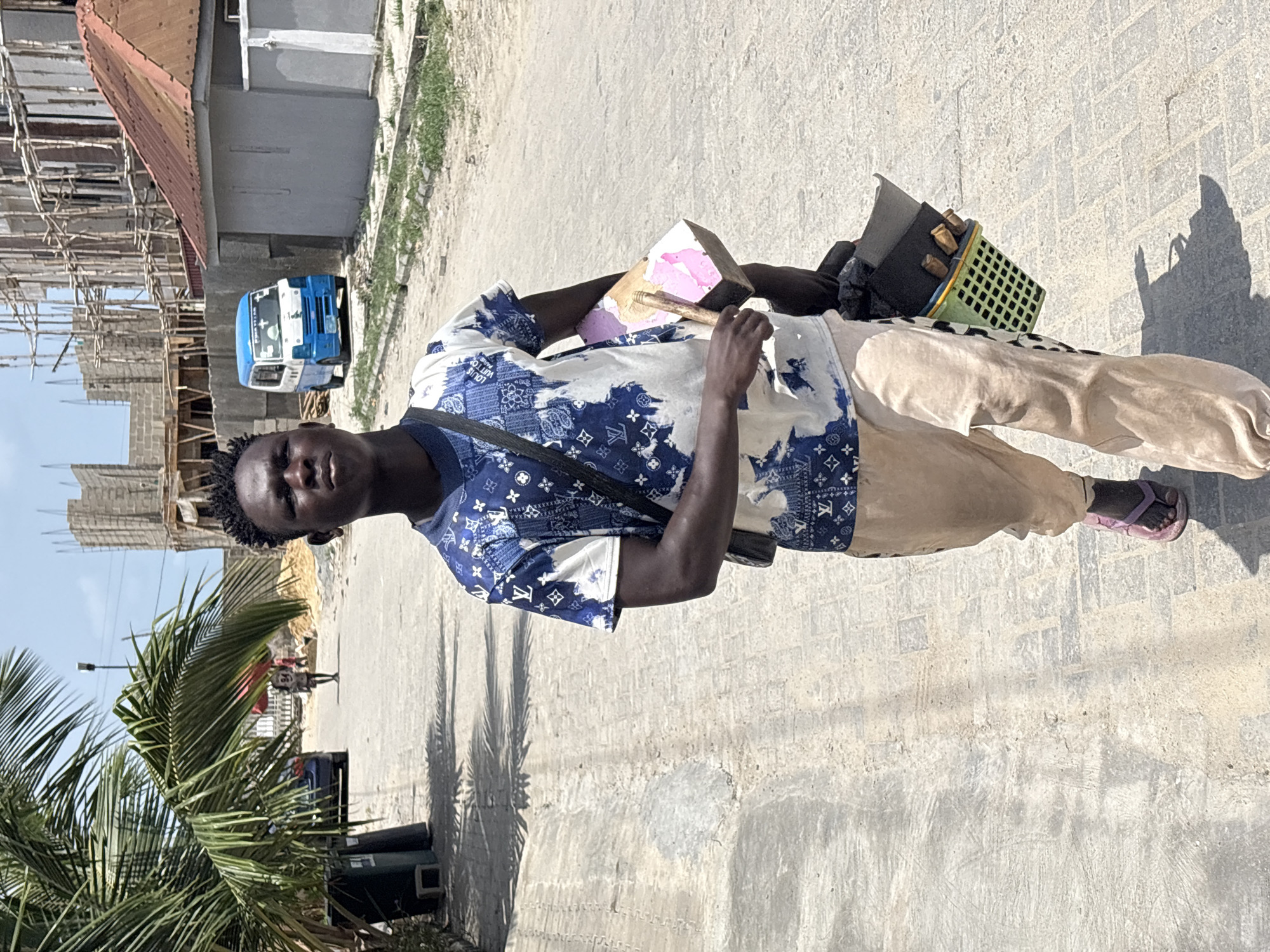 The image captures a young Black man standing on a paved road in Igboefon, Nigeria, under bright, clear sunlight. He appears to be a street vendor or hawker, looking directly at the camera with a neutral expression.
He is wearing a short-sleeved, collared shirt with a blue and white patterned design, featuring motifs reminiscent of Louis Vuitton monograms and bandana prints. He also wears light beige trousers and pink flip-flops, with a dark strap crossing his chest.
In his left hand, he holds a wooden block adorned with a pink substance, possibly paint or a local snack item, and a small brush or stick. In his right hand, he carries a light green plastic crate filled with several small, dark, rectangular goods, likely items for sale.
The background reveals an active street environment. To the left, an unfinished building with extensive wooden scaffolding and concrete block walls indicates ongoing construction. A small blue and white three-wheeled vehicle, resembling an auto-rickshaw, is parked further down the road. A large palm tree with vibrant green fronds is prominent on the right side of the frame, casting long shadows on the light-colored, paved road. The bright sun suggests it is midday or early afternoon.