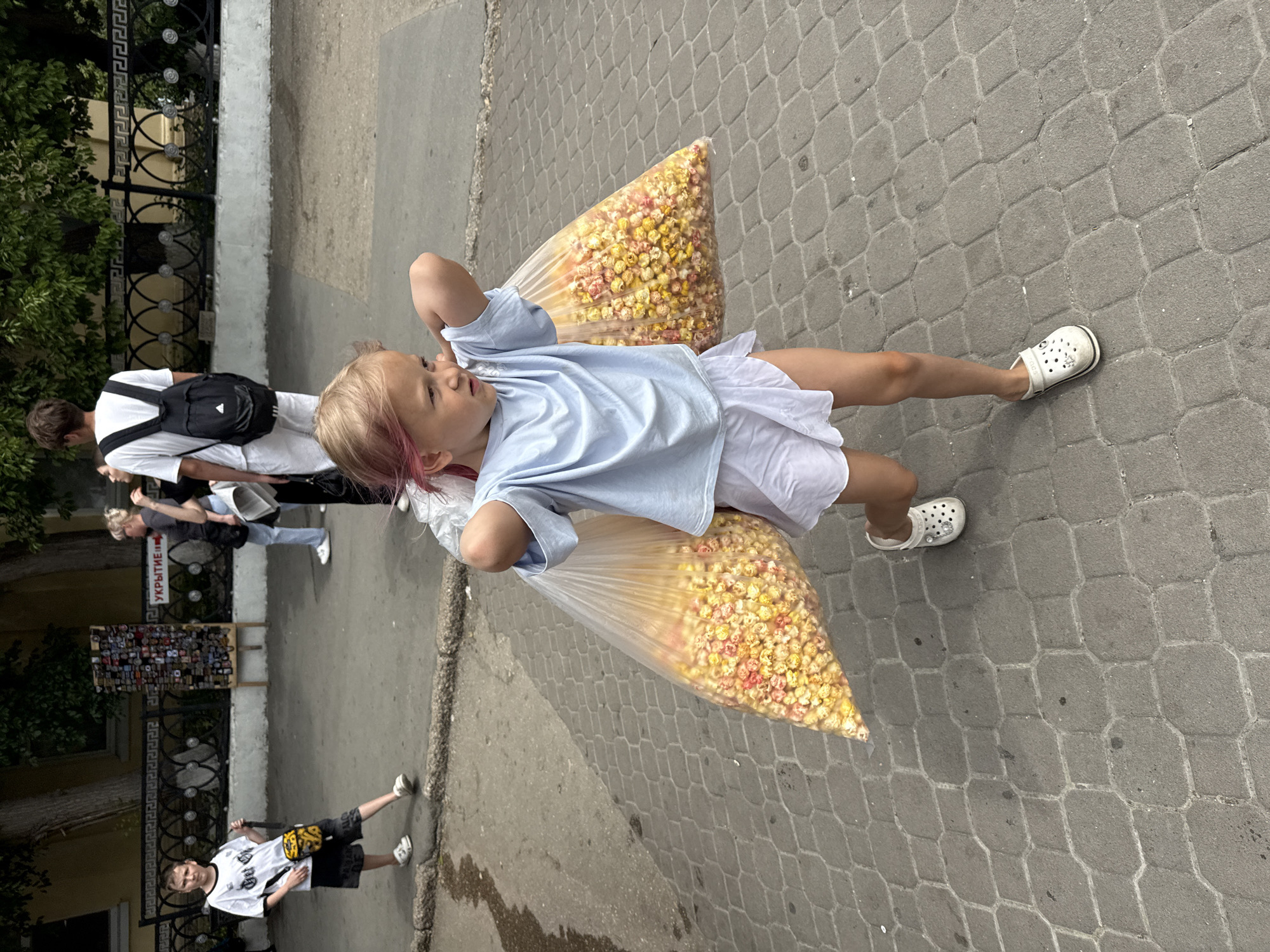 A young blonde girl with pink-tipped hair, wearing a light blue t-shirt and white shorts, walks forward on a gray hexagonal paved sidewalk. She carries two large clear plastic bags filled with multicolored popcorn (yellow, red, white) slung over her shoulders and held with her hands above her head, appearing to struggle slightly with the weight. She wears white Crocs.
In the background, on the left, a boy in a white and black t-shirt and dark shorts, holding a black strap, stands on the sidewalk, looking towards the viewer. Behind him is a metal fence with decorative patterns and an old, light yellow building with trees. To the boy's right, a wooden display stand holds numerous small merchandise items. A red and white sign pointing right reads "УКРЫТИЕ" (SHELTER). Further back, several adults are visible, one wearing a white t-shirt and black backpack, another in a gray top and light blue jeans. The scene is outdoors on a sunny day.
Location: Unknown, Unknown