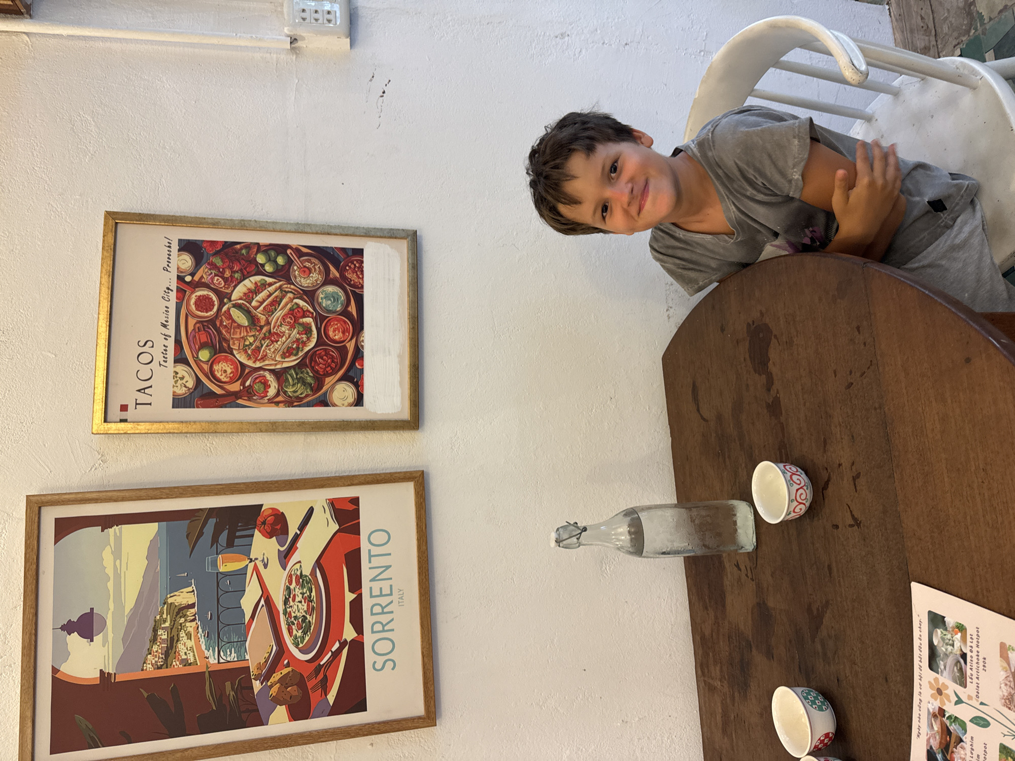 A young boy is sitting at a wooden table in a restaurant. He is wearing a grey t-shirt and grey sweatpants. He has dark brown hair and is smiling at the camera. On the table, there is a bottle of water, two small bowls, and a menu. In the background, there are two framed posters on the wall. One poster is of Sorrento, Italy, and the other is of tacos. The restaurant appears to be casual and brightly lit. The overall scene suggests a pleasant meal with a child.