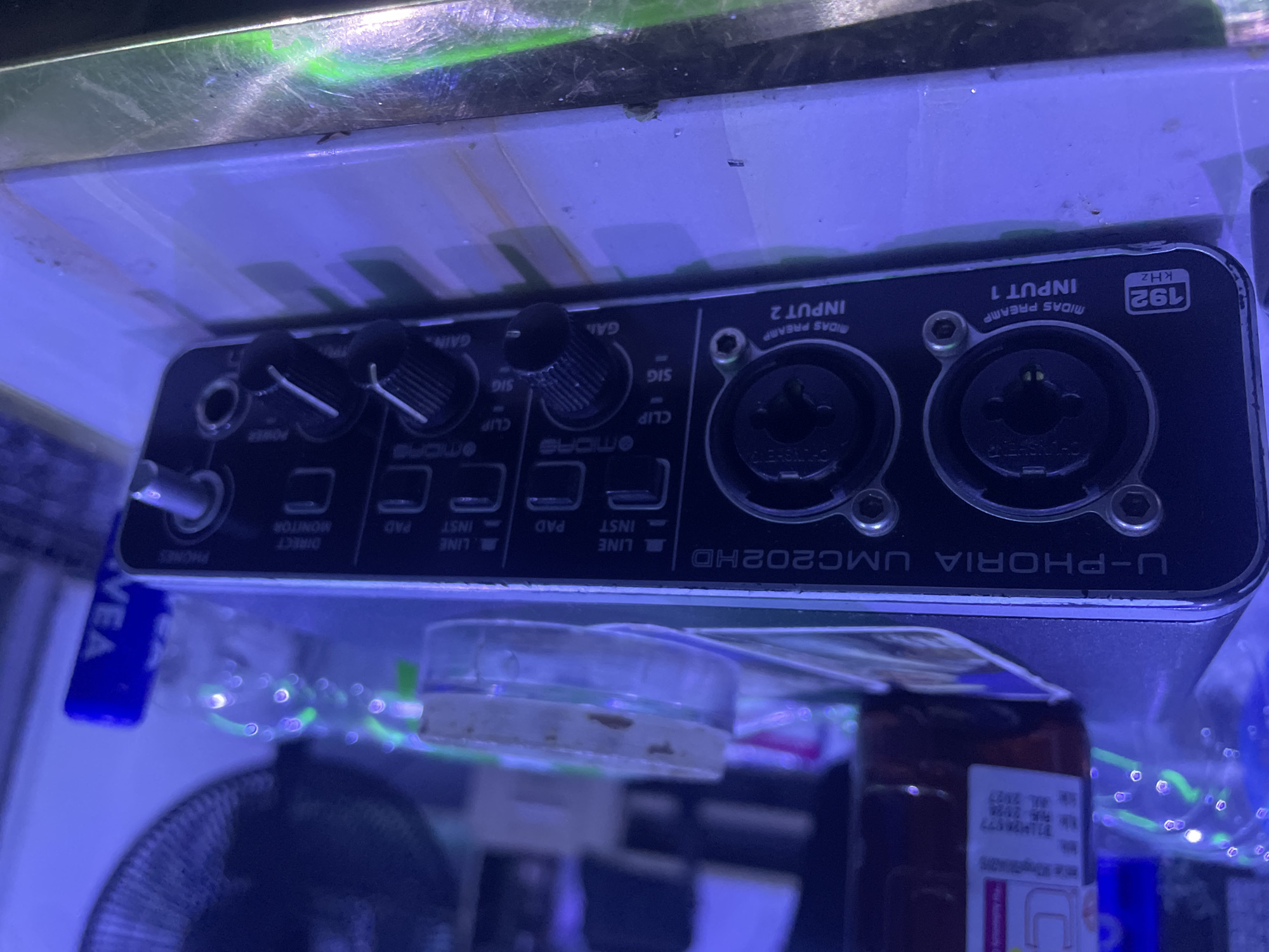 The image displays a close-up, slightly angled view of a Behringer U-PHORIA UMC202HD audio interface, which is the primary subject. The device features two input channels, labeled "INPUT 1" and "INPUT 2," both equipped with "MIDAS PREAMP." Visible controls include two "GAIN" knobs, "PHONES" output, "DIRECT MONITOR" switch, and per-channel "PAD," "INST," and "LINE" selectors. Indicators for "SIG" (signal) and "CLIP" are also present above each input. A "POWER" switch or indicator is on the left, and a "192 KHZ" label is visible on the top right.
No human activity is taking place; the image captures the electronic equipment in a static setup. The scene is illuminated by a predominant blue or purple light, which creates green light reflections on the metallic surface directly above the audio interface. Below the device, parts of a fan, a small clear container, and a brown bottle with a label are visible, indicating an indoor, potentially cluttered environment. A blue object with the partial text "VEA" is seen on the left. The visible text on the audio interface includes "U-PHORIA UMC202HD," "MIDAS PREAMP," "INPUT 1," "INPUT 2," "192 KHZ," "GAIN," "SIG," "CLIP," "POWER," "PHONES," "DIRECT MONITOR," "PAD," "INST," and "LINE." This setup is located in Abuja, Nigeria.