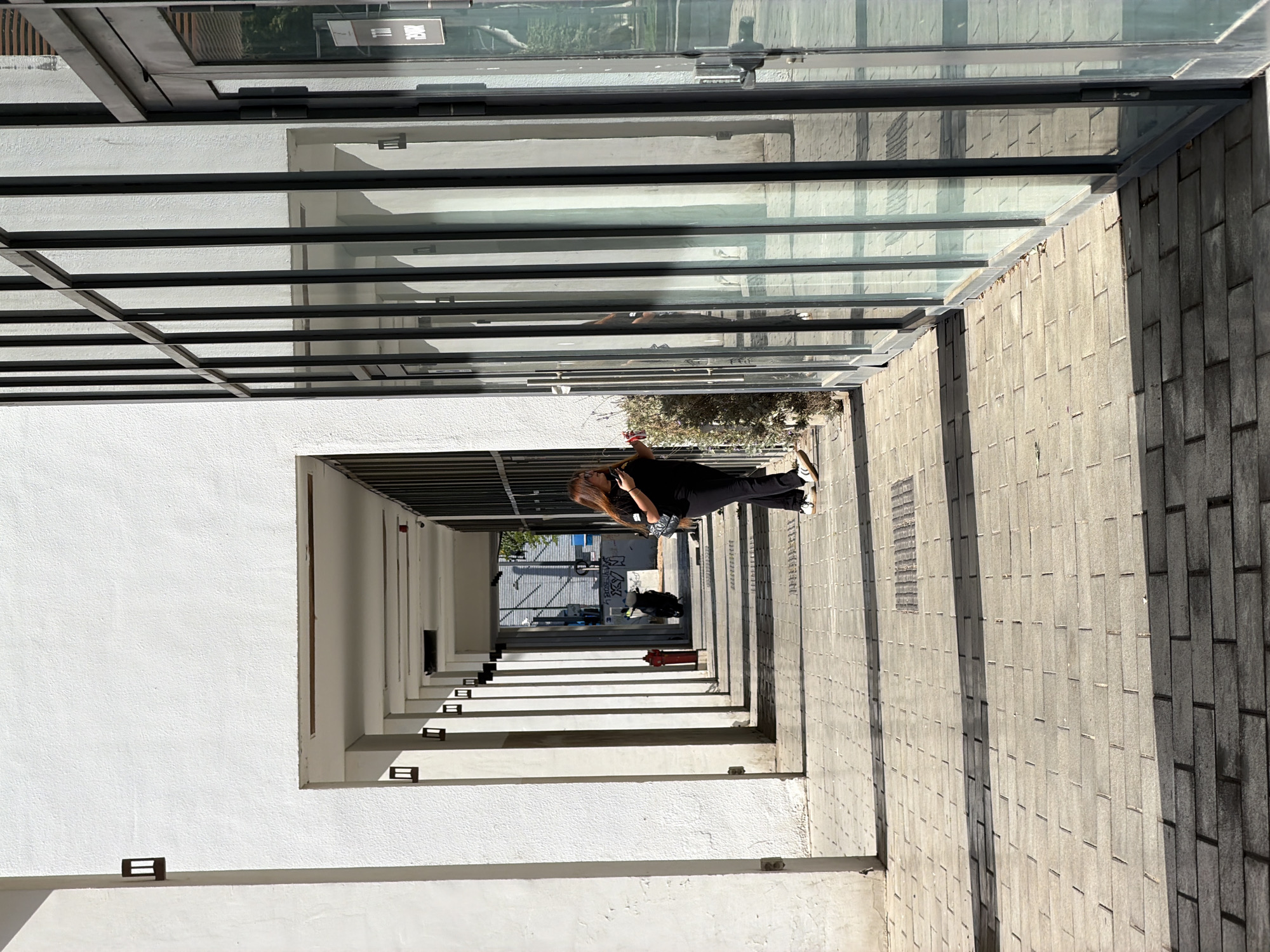 A low-angle shot of a pedestrian passageway located in Tel Aviv, Israel. A woman stands on the gray brick pavement, talking on her phone. A glass and metal awning covers the right side of the passageway, creating a shadow on the pavement. On the left, white pillars create a series of archways. In the distance, the passageway opens to reveal a street scene with cars and buildings. The light suggests it is a sunny day.