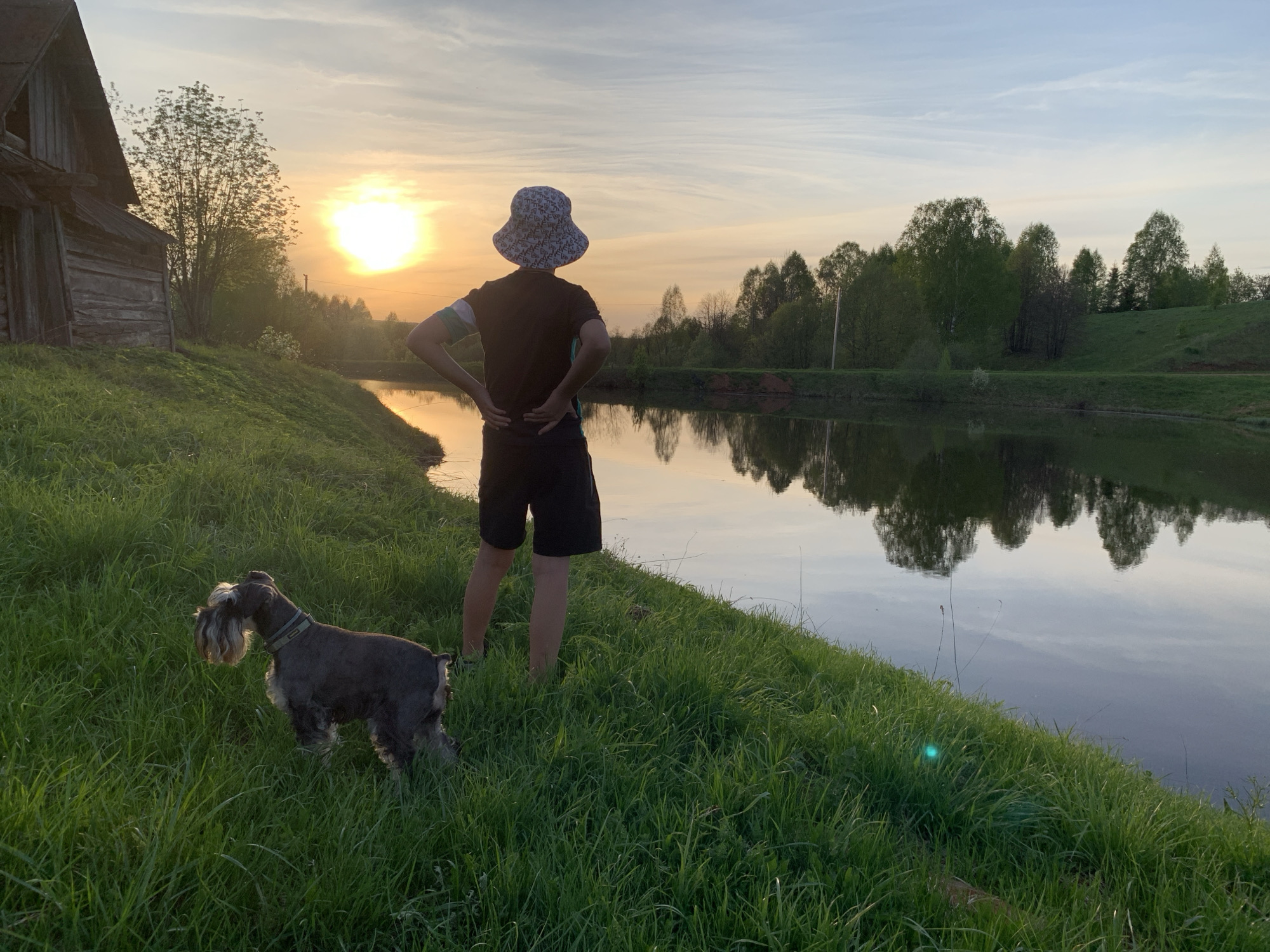 A person in a dark shirt and patterned hat stands on a grassy bank next to a body of water. They are facing away from the viewer with their hands on their lower back. A dog stands in the grass to the left of the person. The water reflects the sky and trees on the opposite bank. A wooden building is visible on the left side of the image. The sun is setting or rising in the background, casting a warm glow. The scene appears to be located in Zasechnyi, Russia.