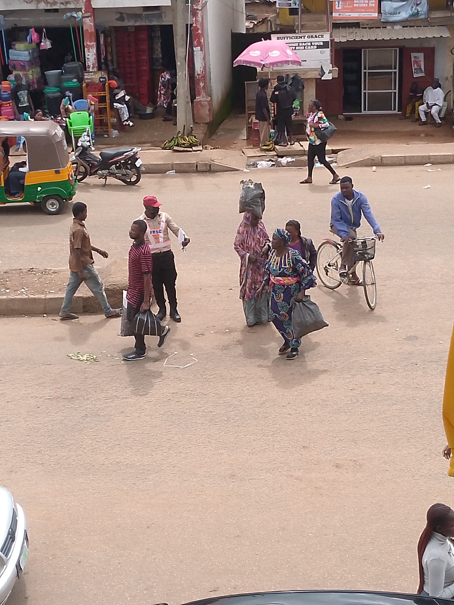 The image captures a bustling street scene in Bukuru, Nigeria. In the foreground, a young boy in a brown shirt and blue jeans walks to the left, while a man in a red cap and a reflective vest, identified by the "FRSC" logo, appears to be directing traffic or speaking to another man in a striped shirt who is carrying a black bag. A motorcycle is parked near a green and yellow tricycle, and in the background, several people are seen near shops. One woman in a colorful dress walks with a large black bag, and a man on a bicycle rides away from the camera. Several bags and other items are scattered on the ground, and there are piles of bananas near a storefront. The overall impression is one of everyday urban life, with a mix of pedestrians, vehicles, and commercial activity.