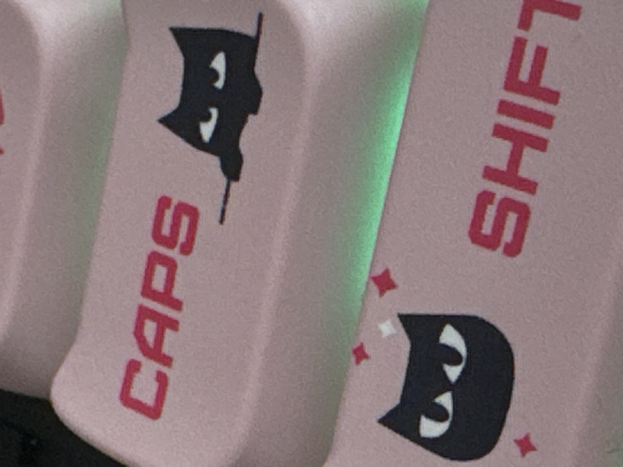 The image is a close-up shot of a keyboard with pink keycaps. The keycaps have red lettering and black cartoon cat designs. One keycap, labeled "CAPS," features a black cat's head peeking over the top, with white eyes. Another keycap, labeled "SHIFT," also has a black cat's head with white eyes, and small red and white star-like shapes are scattered around it. A green light source is visible to the right of the "SHIFT" key. The image is well-lit and shows the textured surface of the keycaps. The overall aesthetic is cute and playful. There are no people or distinct environmental features visible, making it difficult to infer a specific time of day or weather conditions. The location context of Mikhaylovsk, Russia, does not provide any visual cues within the image itself.