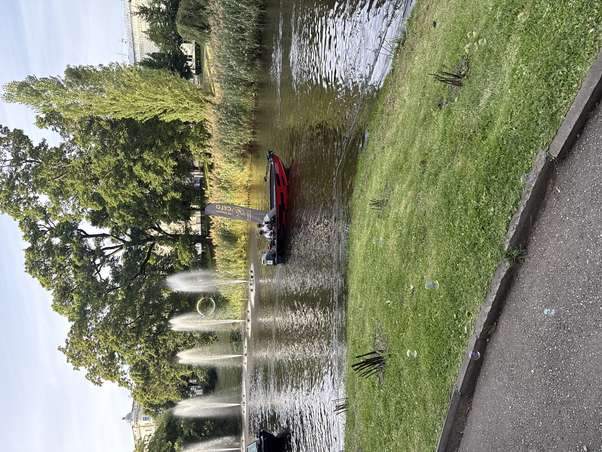 The image depicts a canal in Riga, Latvia, with a small red and black motorboat carrying two people in it. The boat has a flag attached to it. The canal is surrounded by lush green trees on the left and a dense growth of reeds on the right. In the background, there are water fountains spraying water upwards. The sky is partly cloudy and the sun is shining. There are several bubbles floating in the air and on the grass in the foreground. A paved path runs along the edge of the canal. The overall scene suggests a peaceful and pleasant day in an urban park.