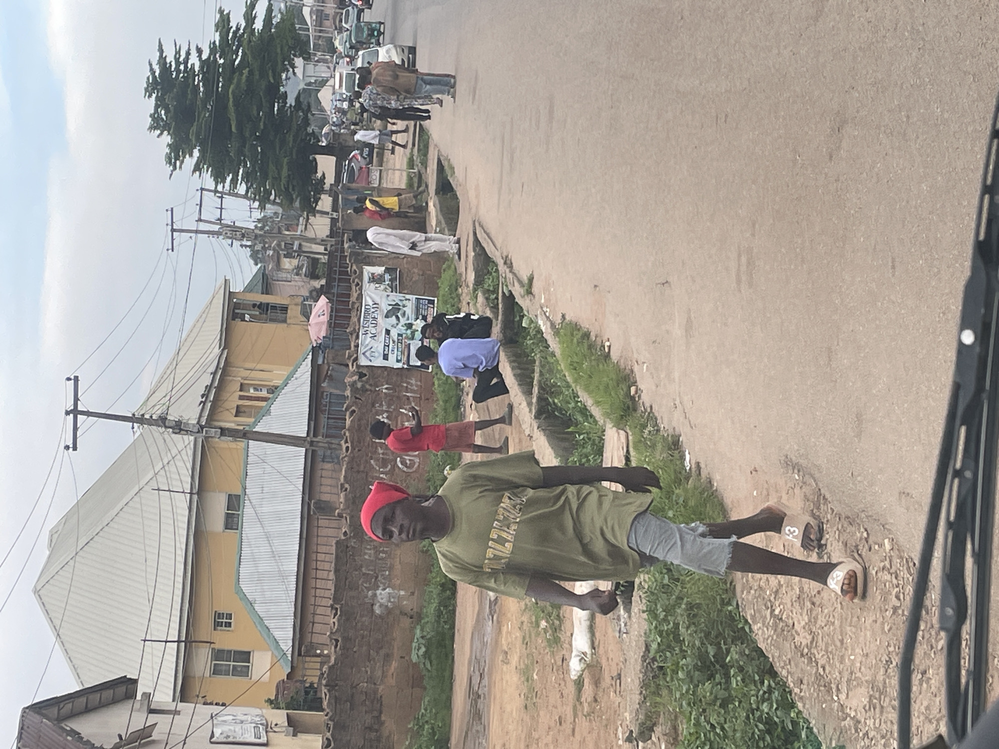 A young male, wearing a red headscarf, an olive green t-shirt with yellow lettering, and light gray denim shorts, walks on a paved shoulder next to a road. He is wearing white slide sandals. He is walking towards the left of the frame. To his right, across a drainage ditch, several people are gathered. Some are sitting on the edge of the ditch, including one person in a blue shirt and another in a dark top. Others are standing, including a man in a white robe, a man in a yellow shirt, and a man in a brown jacket. Behind them, a wall with graffiti stands, topped by a metal fence. A sign for "WISBIRD ACADEMY" is visible on the wall. Further down the road, traffic is moving, with several cars and a tricycle visible. The environment appears to be an urban setting in Jos, Nigeria, characterized by the buildings, road infrastructure, and the presence of various vehicles and pedestrians. The sky is overcast.