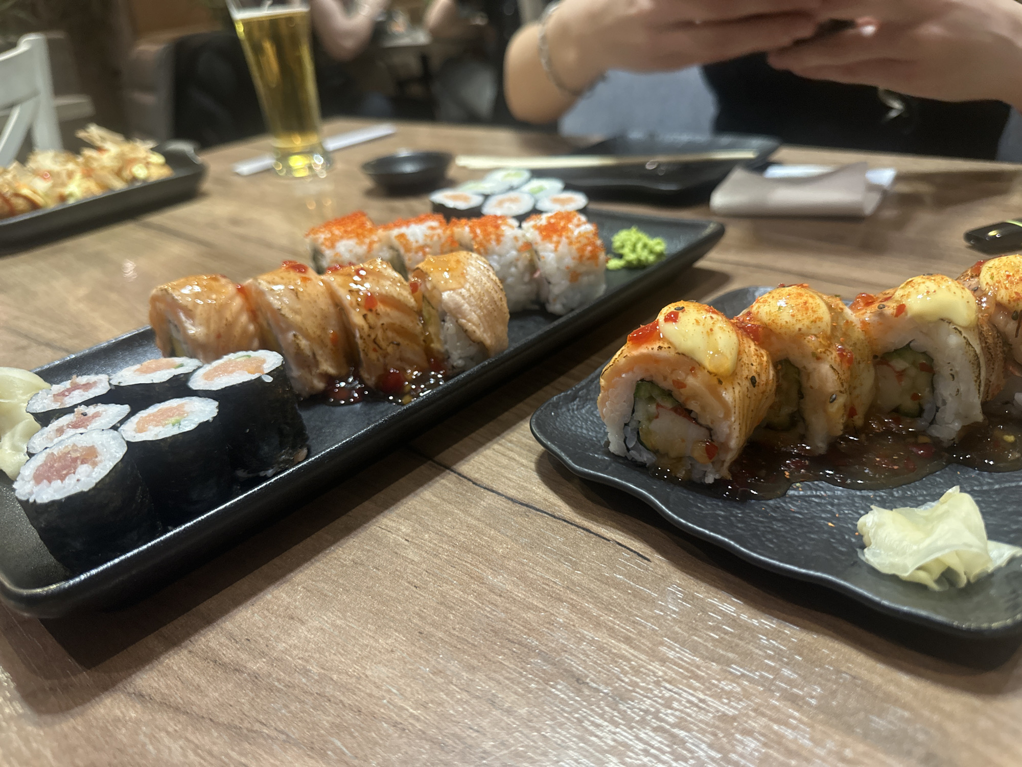 The image shows a table in a restaurant, featuring several plates of sushi. On the left, there is a plate with maki rolls and seared salmon sushi, covered in a dark sauce. Another plate displays rolls topped with spicy mayo. Further in the background, a plate of tempura and a beer glass can be seen. Two people are partially visible in the background, one of them using their phone. The scene likely takes place at a sushi restaurant, possibly in Varna, Bulgaria.
