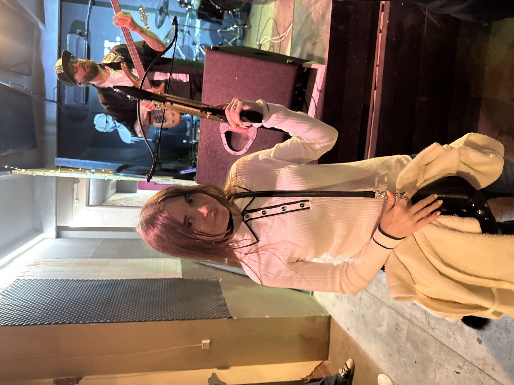 A woman wearing a white, long-sleeved top with black trim is holding a crossbow in front of a dimly lit stage area. A person with a beard and a baseball cap is visible behind her, playing a bass guitar. The background includes stage equipment and a band setup. The woman is also wearing a light-colored coat draped over her arm. The location appears to be indoors with a wall featuring a patterned surface on the left and concrete flooring. The location is Chaikovsky, Russia.