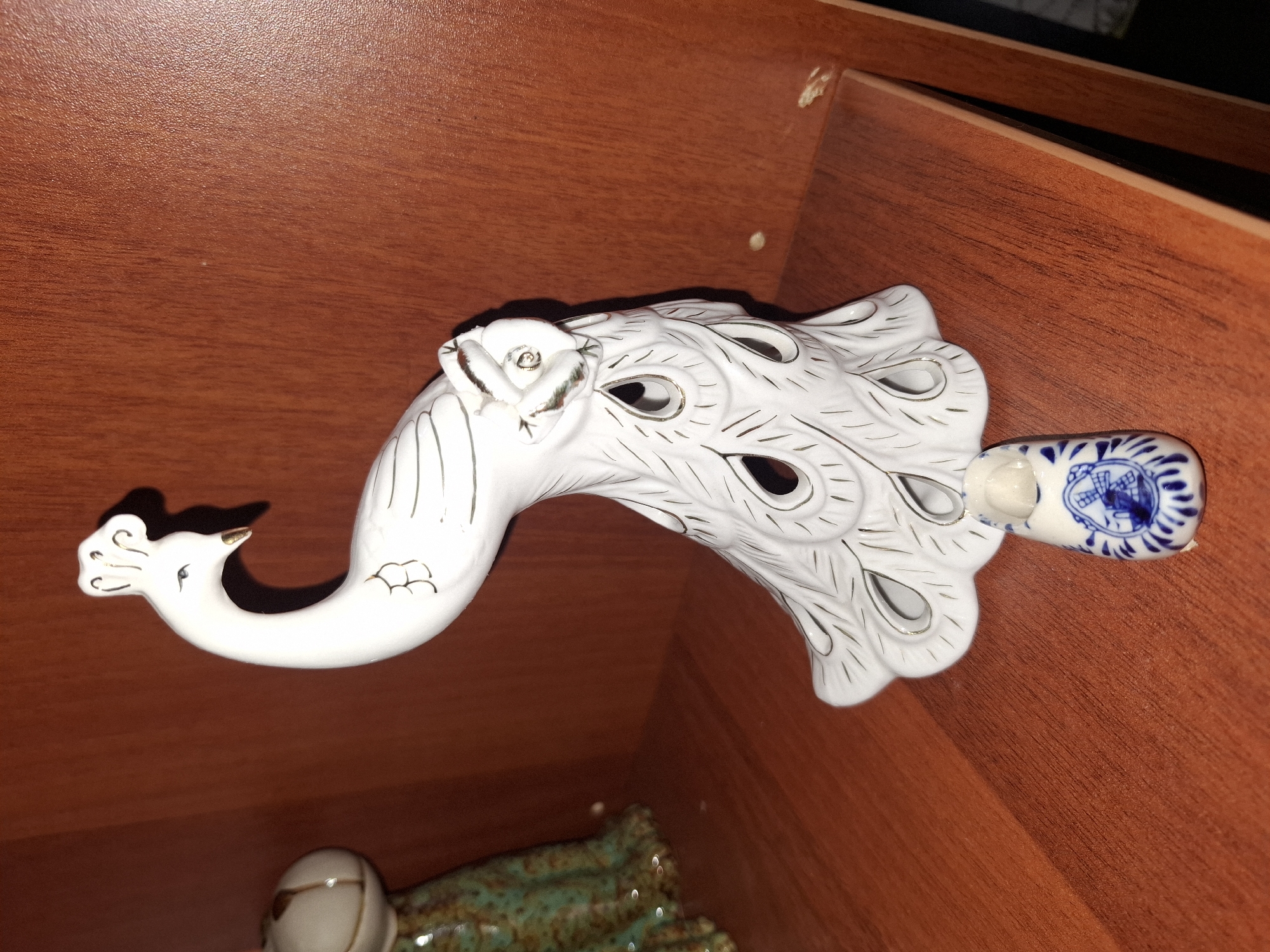 The image shows a white ceramic peacock figurine with gold accents, positioned on a wooden shelf. In front of the peacock is a small ceramic shoe, painted blue and white with a windmill design. The shelf is made of wood with a reddish-brown hue and appears to be part of a larger piece of furniture, possibly a bookshelf or display cabinet. The background is a darker wooden surface. There is a hint of another decorative object to the left of the peacock. The lighting suggests an indoor setting, likely a home or a shop. The image does not provide any information about the city of Almaty, Kazakhstan.
