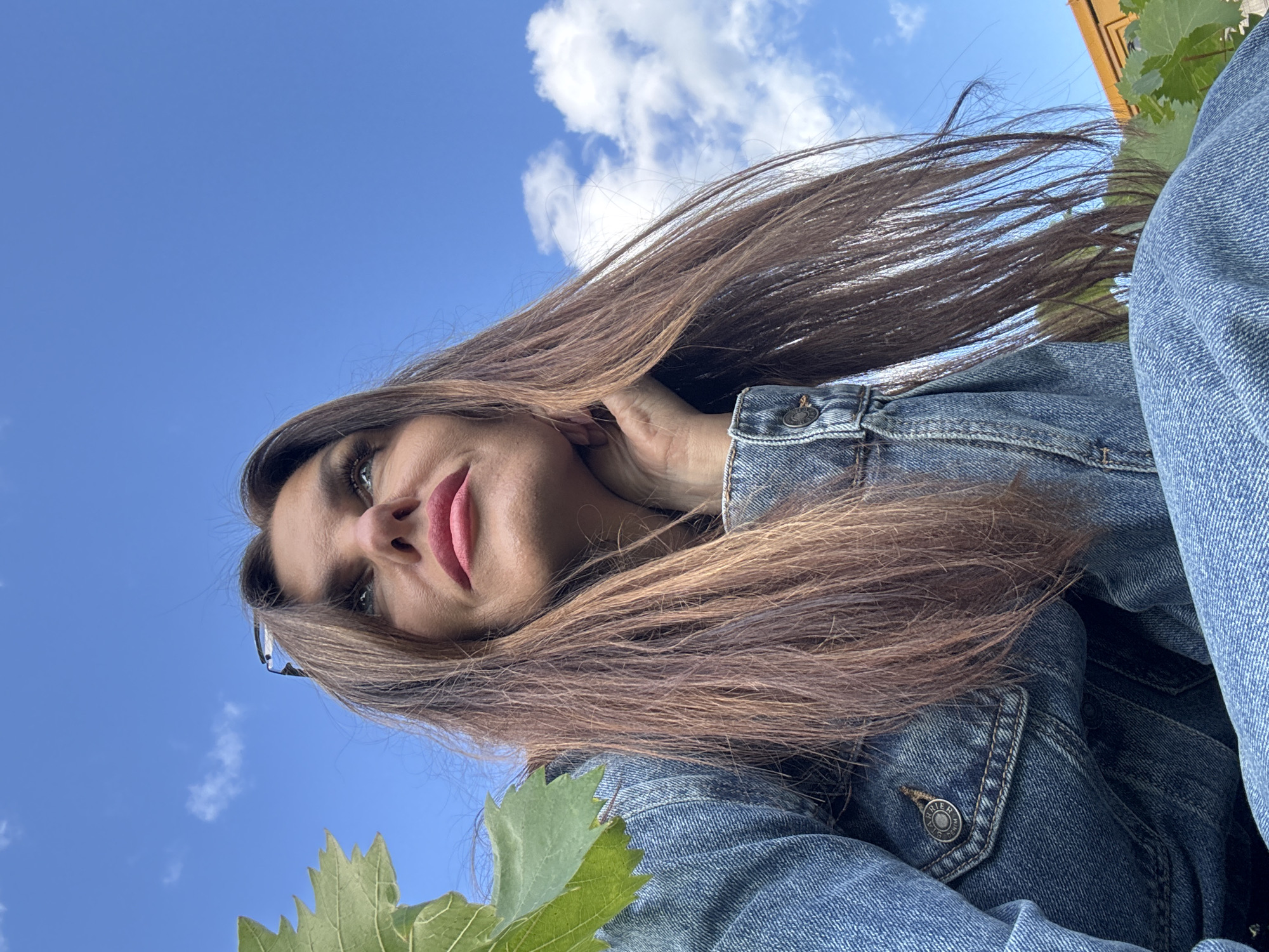 A low-angle shot features a woman with long, brown hair wearing a denim jacket, looking up at a bright blue sky with scattered clouds. She is resting her chin on her hand. A green leaf is visible in the foreground on the left. In the background, a portion of an orange building is visible, along with some greenery and a hint of a treeline. The scene appears to be outdoors, possibly in a rural or semi-rural setting, and does not contain any specific identifying features of Dubovaya, Russia.