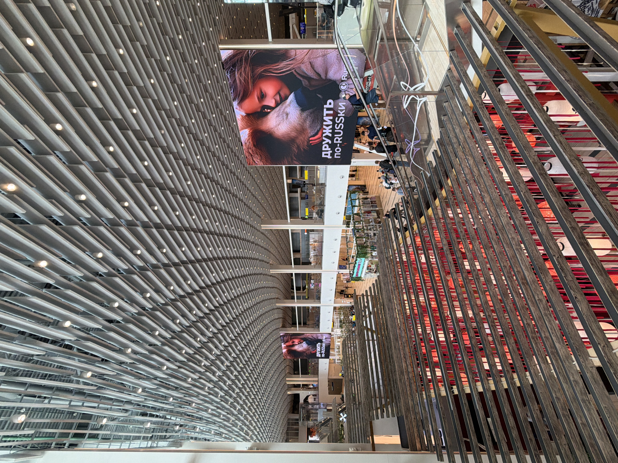 This image shows the interior of a large, modern building, likely an airport or a shopping mall, with a high, slatted ceiling and multiple levels. In the foreground, there are rows of wooden beams and metal railings, suggesting balconies or walkways overlooking a central area. Below, red and white fabric or decorations hang, with some white lanterns visible.
Dominating the scene is a large digital billboard displaying an advertisement. The advertisement features a close-up image of a young girl with blonde hair embracing a bear. Below the image, text in Russian and English reads "ДРУЖИТЬ по-RUSSKi" which translates to "Befriend the Russian Way." A logo that appears to be for Ru.Stock is also visible.
Several people are visible in the image, walking on escalators, sitting on benches, and browsing in shops on the lower levels. The overall environment appears clean, well-lit, and contemporary. The image is taken from an elevated perspective, looking down into the main concourse of the building.
The advertisement and the text "по-RUSSKi" strongly suggest a connection to Russia. Without more specific visual cues, it is difficult to pinpoint the exact location in Yuzhno-Sakhalinsk, Russia, but the scale and modern amenities indicate it could be a major public space such as a transportation hub or a large commercial center.