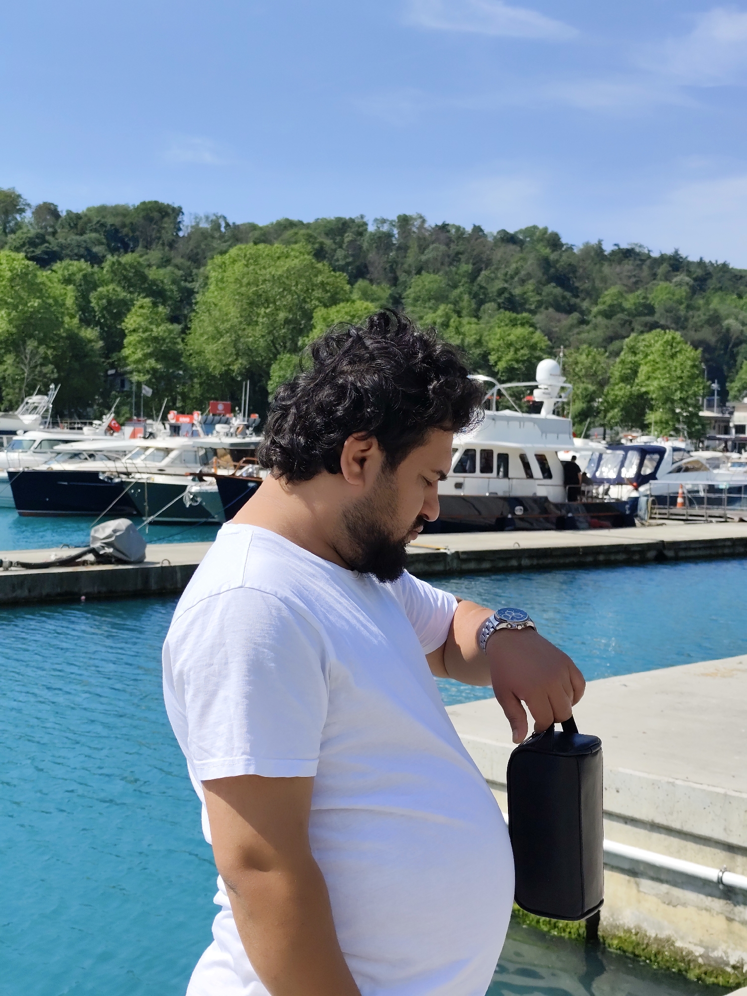 A man stands on a concrete dock in Sarıyer, Turkey, with turquoise water and boats in the background. The man has dark curly hair, a beard, and wears a white T-shirt. He's holding a black cylindrical bag in his right hand and looking at his wristwatch. Behind him are parked boats and a tree-covered hillside, under a clear sky.