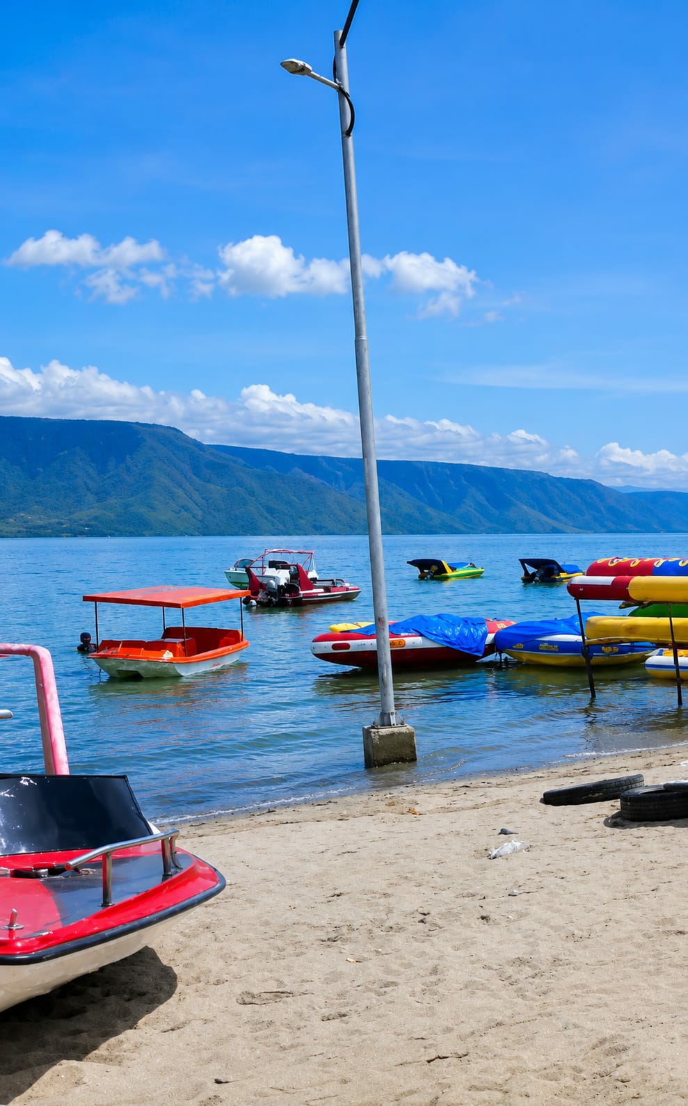 The image depicts a sunny day at the beach in Situngkir, Indonesia. The foreground features a sandy beach with two red and white boats resting on the shore, one with a black seat. In the shallow water, a pink structure is visible on the left, and a small orange boat with a canopy is anchored. Further out in the bright blue water, several colorful boats are floating, including a red and white motorboat with an outboard engine, and two yellow and green pedal boats. The background is dominated by a large, blue mountain range under a clear blue sky with scattered white clouds. A tall, grey lamppost stands in the middle ground, casting a shadow on the sand. The overall scene conveys a sense of a peaceful and recreational lakeside environment.