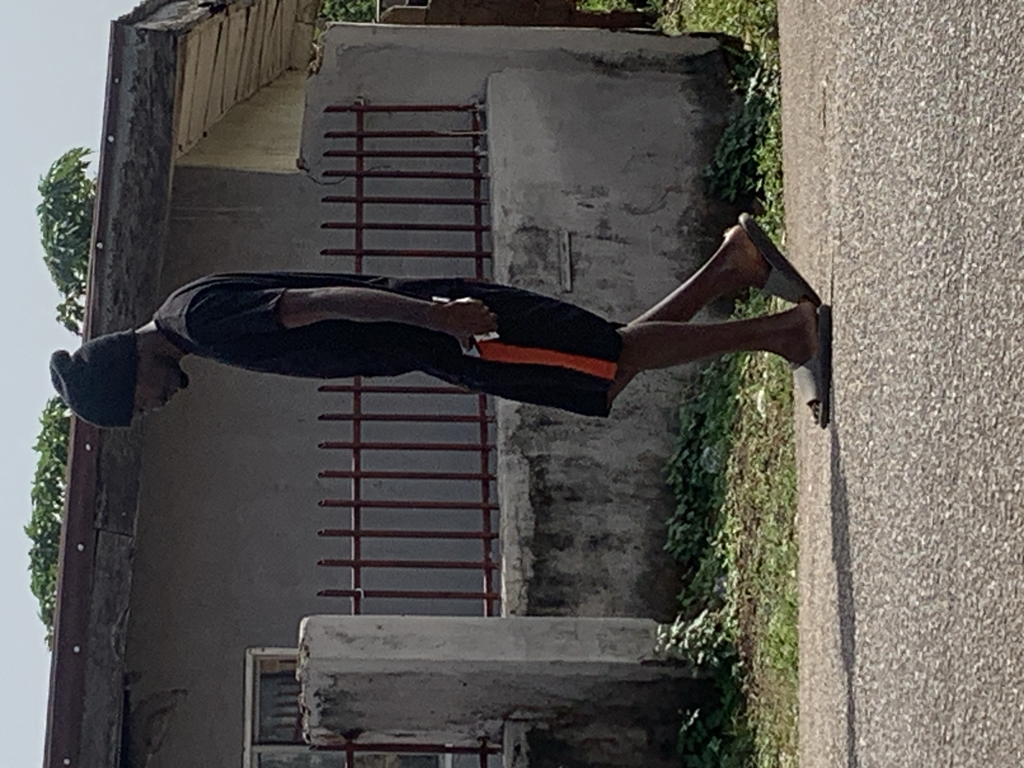 The image depicts a man walking along a paved road in Gura Top, Nigeria. He is wearing a black beanie, a black t-shirt, and black shorts with an orange stripe down the side. The man is holding a white object, possibly a phone, in his right hand. He is wearing grey flip-flops. In the background, there is a concrete wall with a red metal fence in front of it. Behind the fence, a building with a damaged roof and a window is visible. Green foliage grows along the base of the wall. The lighting suggests it is daytime.
