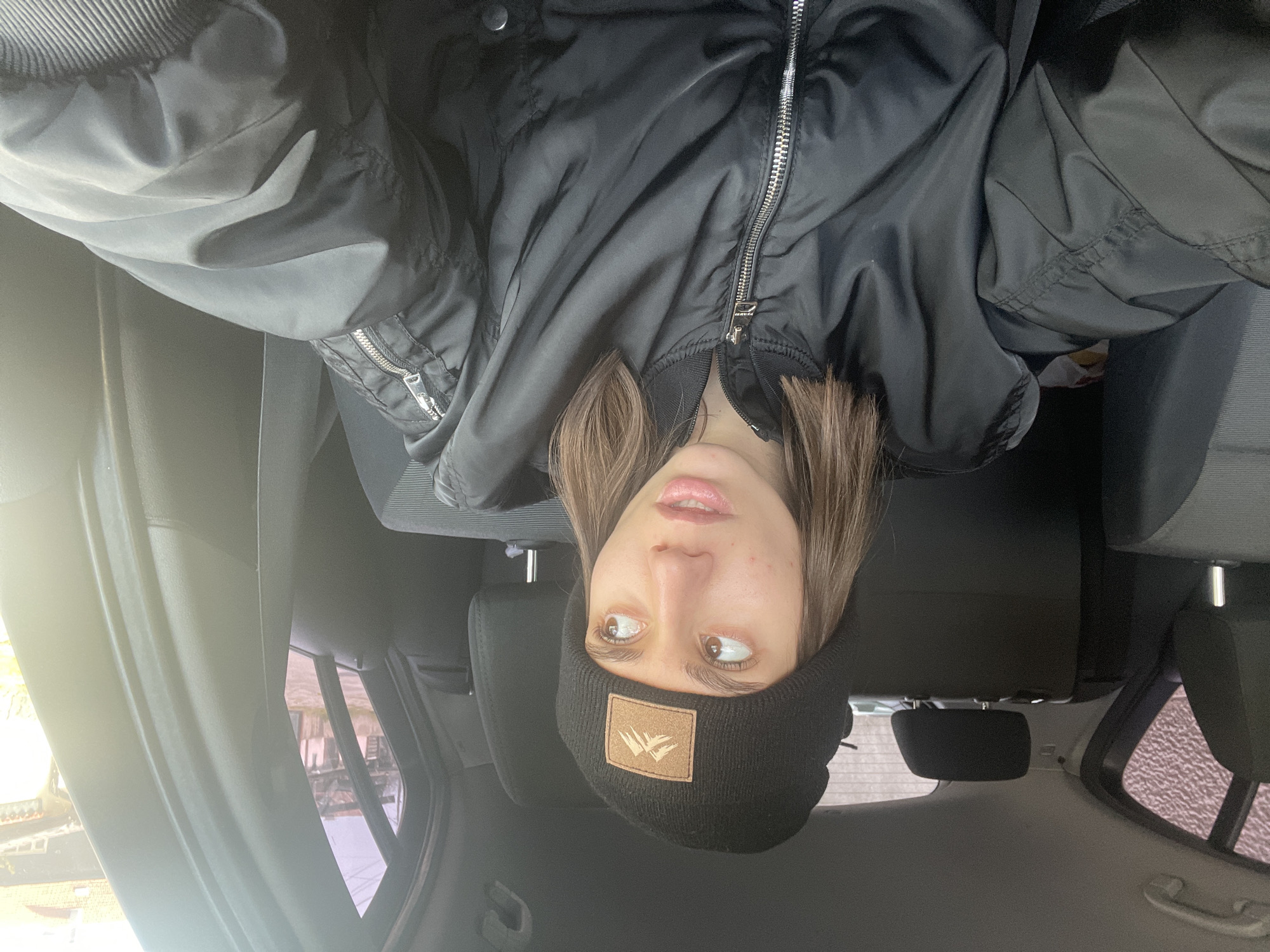 A young woman is in the backseat of a car. She wears a black bomber jacket and a black beanie with a tan patch. The interior of the car is gray, with adjustable headrests and a hand grip on the ceiling. The window shows a view of a building outside, blurry due to the bright light. The location cannot be determined from this image.