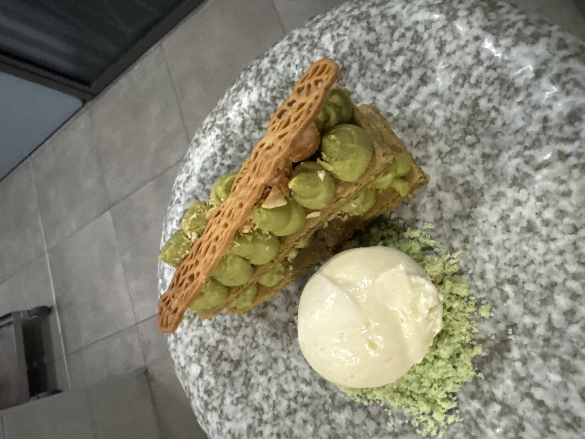 A plated dessert consisting of a mille-feuille filled with green cream and topped with a decorative crisp, served with a scoop of pale ice cream on a bed of green crumble. The dessert rests on a speckled grey plate. In the background, a tiled floor and a metal cart with wheels are visible. The scene appears to be indoors, likely in a restaurant or food establishment. No specific location in Sofia, Bulgaria can be determined from the image.