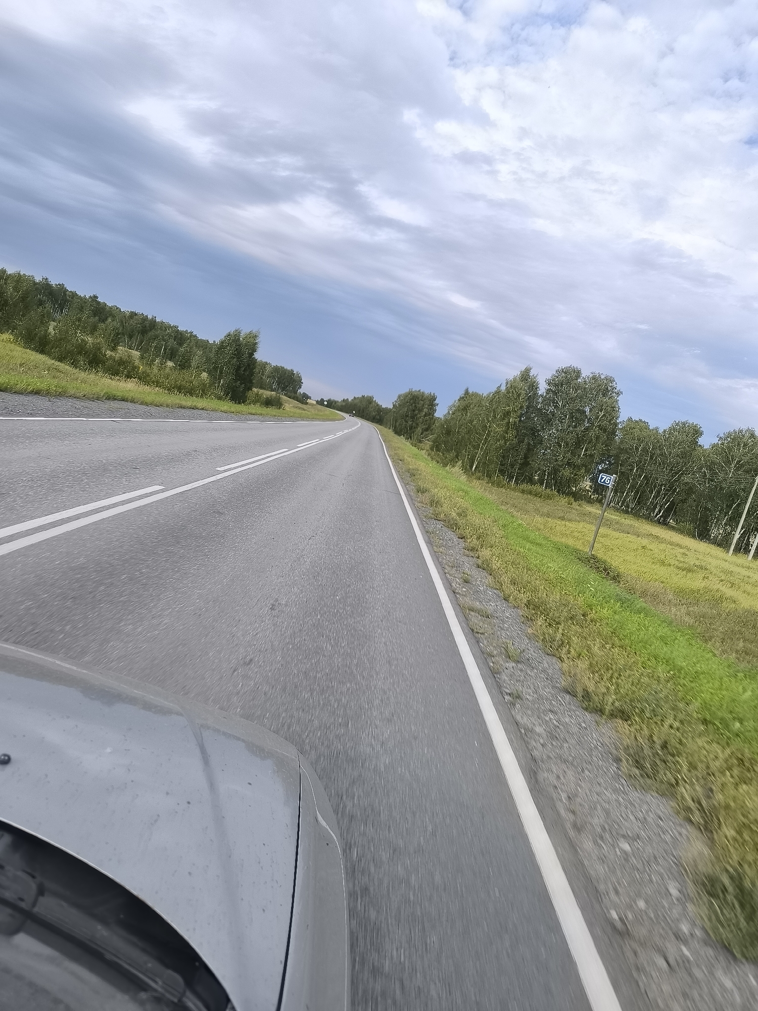 The image depicts a gray asphalt road with double white lines, suggesting it is a two-lane highway. The road curves slightly to the right and appears to be moving away from the viewer. On the right side of the road, there is a grassy embankment with trees beyond. A signpost with the number "76" in a blue circle is visible on the right. The sky is cloudy, with patches of blue visible. The front of a silver car is visible in the lower left corner of the image, indicating the photo was taken from inside a moving vehicle. The scene appears to be in a rural or semi-rural area, with no buildings or significant urban infrastructure visible.