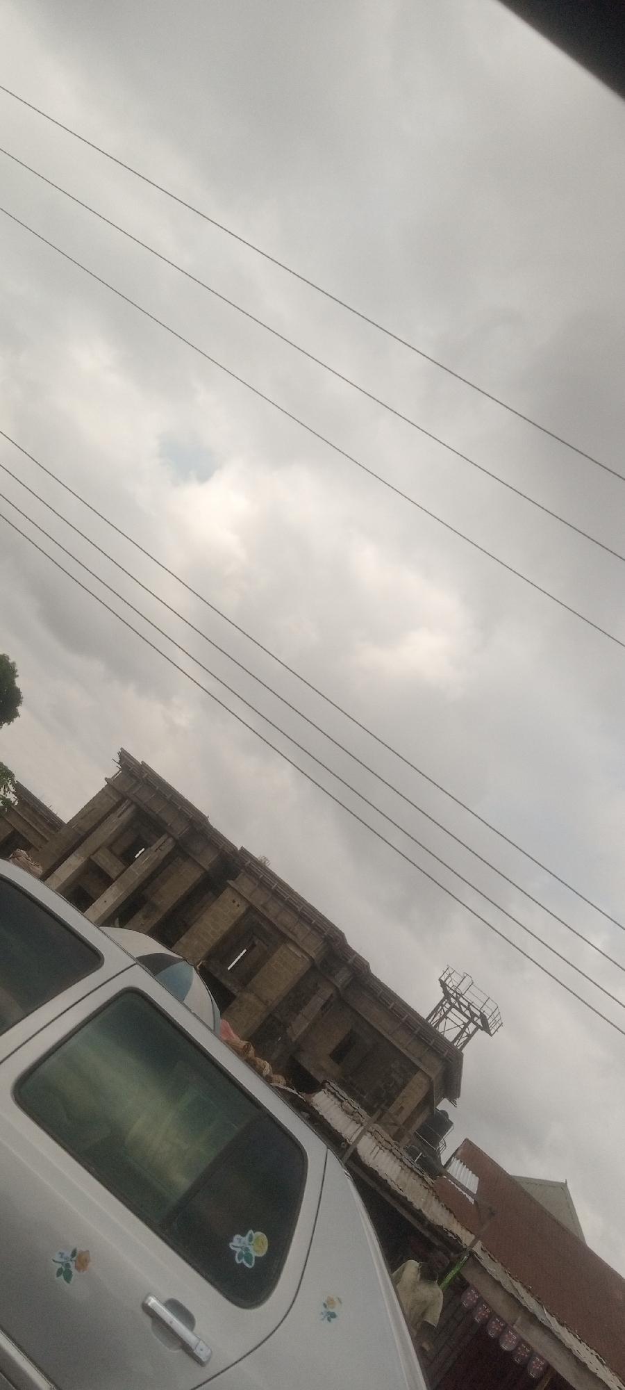 The image, taken in Sabon Tasha, Nigeria, presents a low-angle shot. An off-white vehicle with flower stickers on its side is prominently featured in the foreground. Behind the vehicle, a dilapidated concrete structure with visible framework stands under a cloudy sky. Power lines stretch across the upper portion of the frame. The building appears to be partially constructed or in disrepair, with open window spaces and an observation tower on top. A partially visible red brick building is situated to the right of the concrete structure. The overall tone of the scene is overcast.