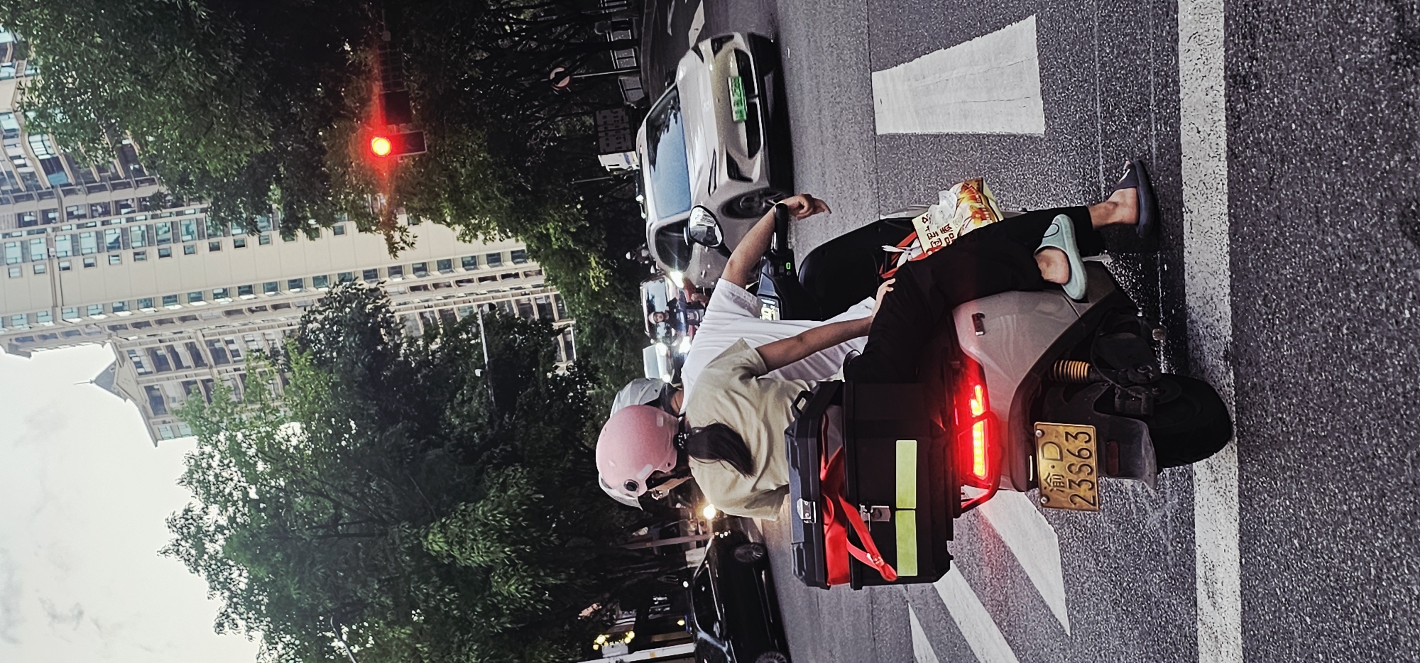 Two people are sitting on a gray scooter. The driver is wearing a pink helmet and a beige shirt. The passenger is wearing a white shirt and black pants with light blue sandals. The scooter has a black box strapped to the back with red straps. The license plate is visible and reads "渝D 23S63". The scooter is stopped on the road at a crosswalk with a red light. There are buildings, trees, and other vehicles in the background. The location is likely in the Yuzhong District of China, based on the "渝" character on the license plate.