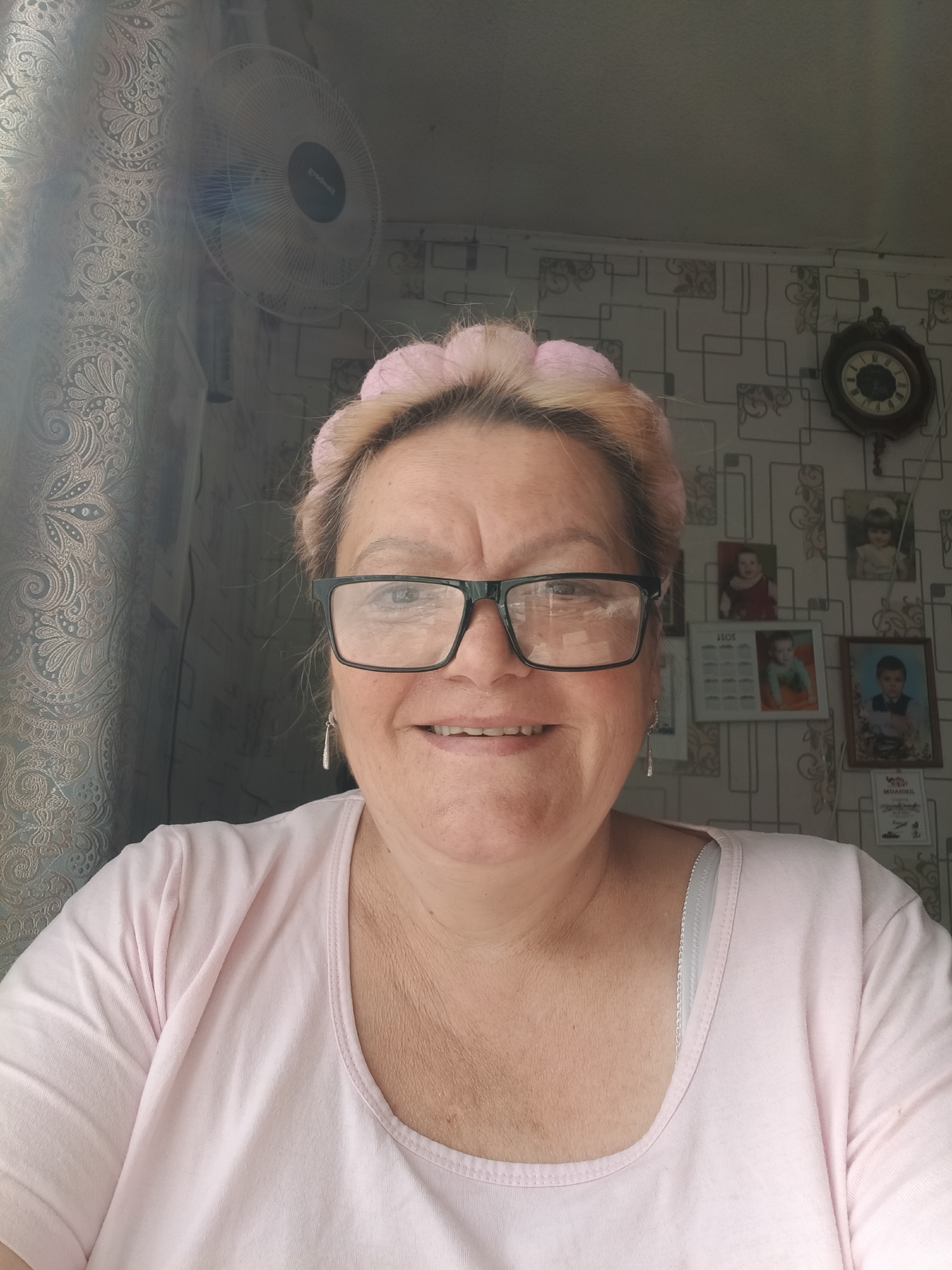 The image depicts a woman in Красный Маяк, Russia. She wears glasses and a light pink headband, and a light pink t-shirt. A standing fan is visible to her left, next to a window. Behind her is a patterned wall with a clock and several framed photographs.