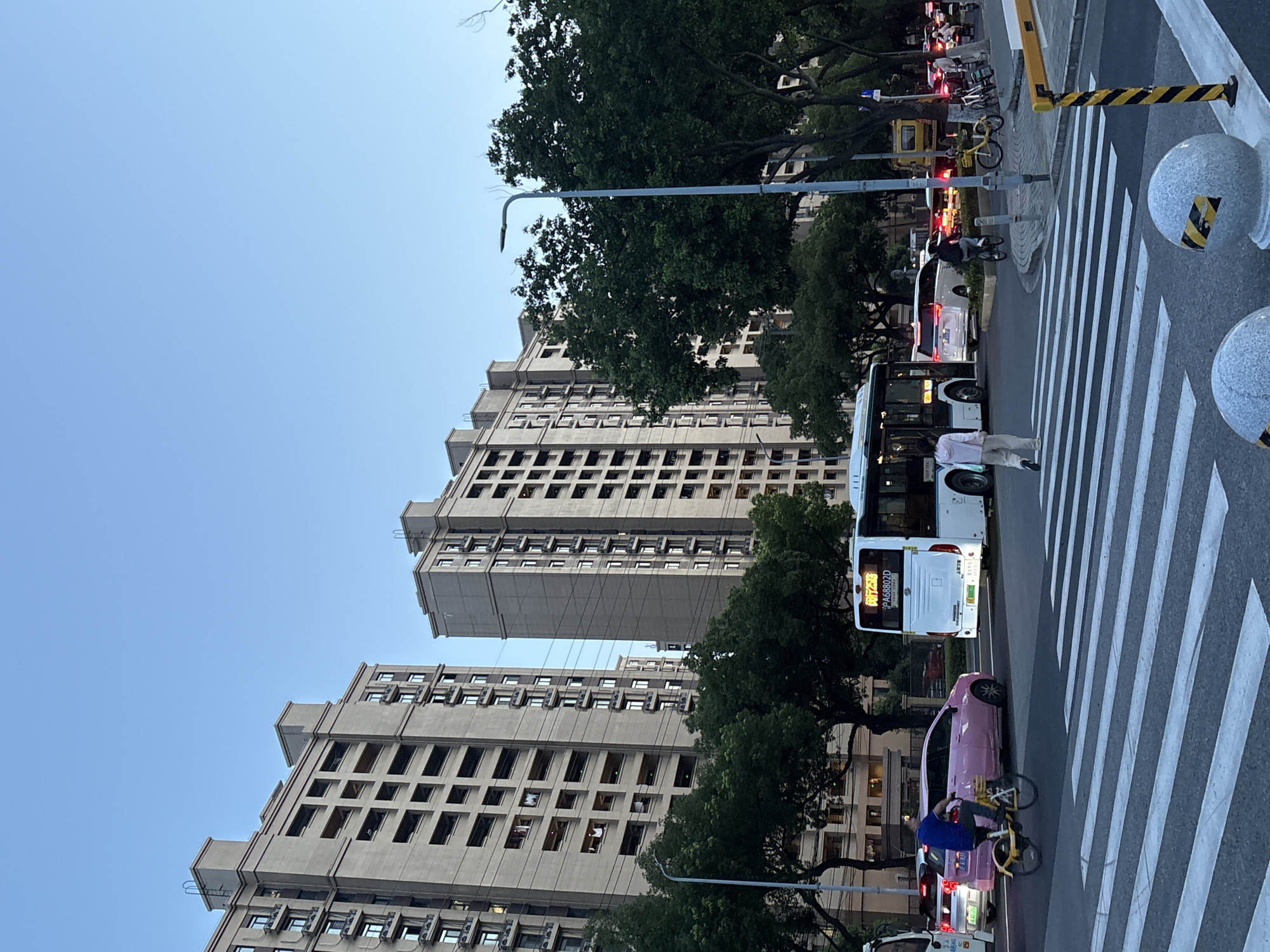 The image shows a street scene in Minhang District, China. Two tall residential buildings are visible in the background, with balconies and many windows. In the foreground, a zebra crossing is in view, with traffic on the road. A white bus is stopped at the crossing, and a person is walking across the street. A pink car is driving past the bus, and a person on a bicycle is also visible. There are trees lining the street and streetlights. In the distance, more cars and buildings are visible. The sky is clear and blue, suggesting it is daytime.