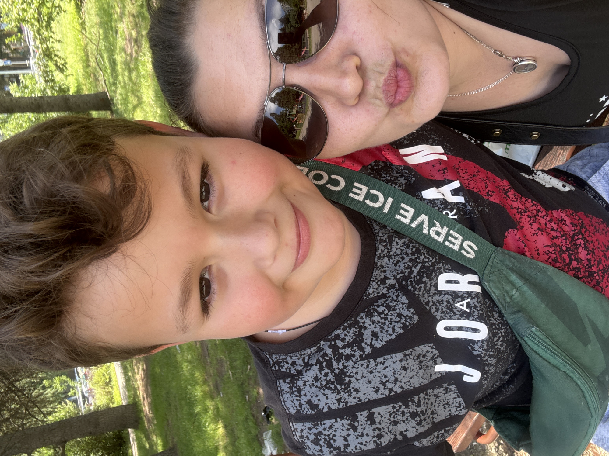 A selfie taken outdoors shows a woman and a young boy. The woman is wearing sunglasses, has dark hair, and is making a kissing face. She is wearing a black top and a silver necklace. The boy has brown hair, brown eyes, and is smiling. He is wearing a black shirt with a speckled gray design, a green fanny pack and a beaded necklace. The background appears to be a grassy park with trees. Reflections of trees and buildings are visible in the woman's sunglasses. The location cannot be determined from the image alone.