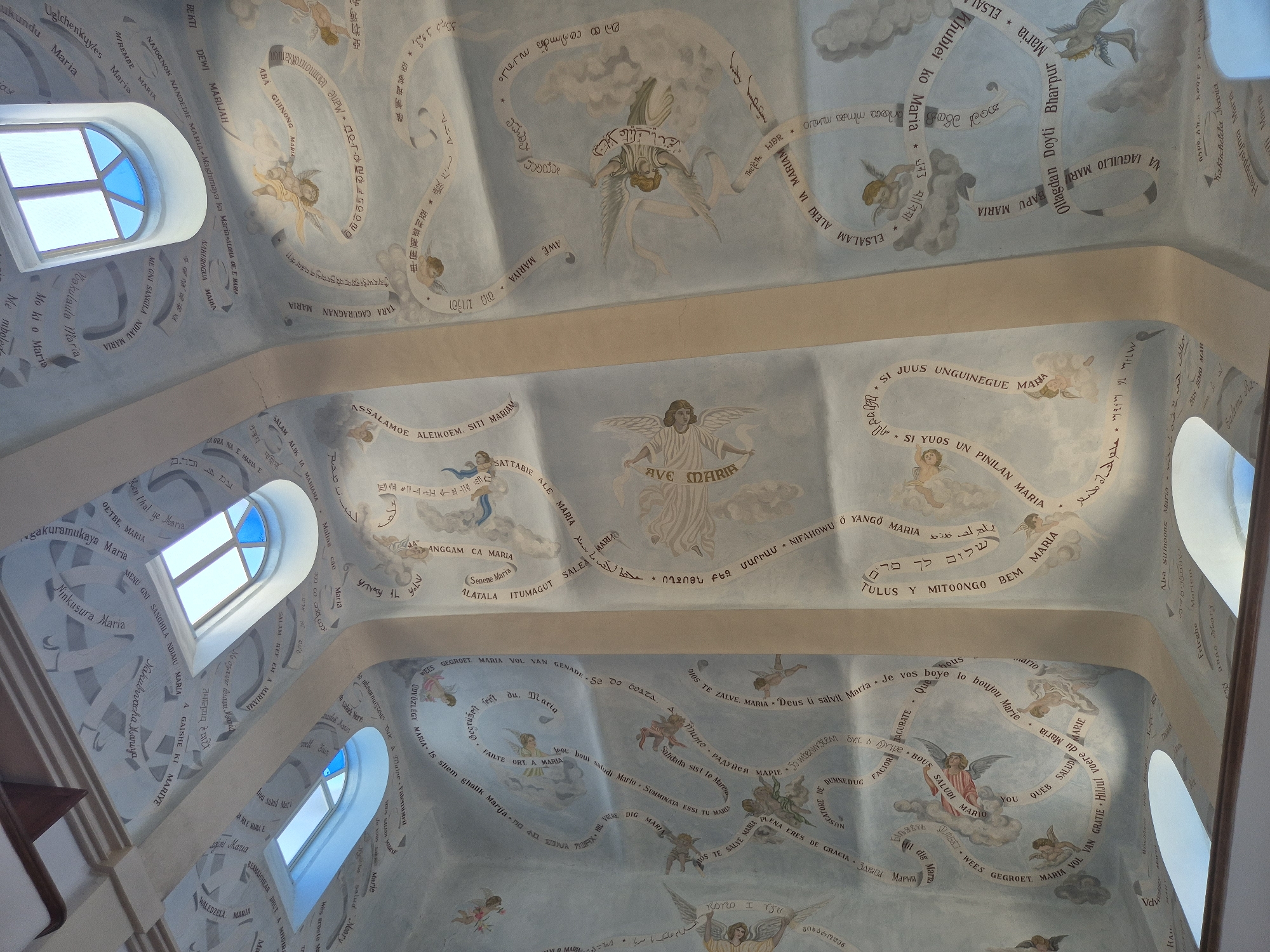 This image depicts the interior of a church or chapel with a painted ceiling adorned with religious imagery and text. The ceiling is curved and features various depictions of angels and cherubs. Scrolls with text are painted along the ceiling, appearing to be prayers or religious inscriptions in multiple languages, including what looks like Hebrew, Arabic, and possibly Latin or other European languages. Two arched windows with blue-tinted glass are visible, letting in natural light. The architecture suggests a traditional religious building. The text and imagery, particularly the depiction of Mary, indicate a Marian shrine or a church dedicated to the Virgin Mary. The question requests location information for the city of Mate Yehuda Regional Council, Israel. While the image itself does not directly indicate this location, such an inscription as "Mishkan Maria" or similar Marian references might be found in churches or shrines within this region of Israel that has a Christian presence. The style of painting and architecture is consistent with religious art found in various parts of the world, but without more specific contextual clues within the image, pinpointing the exact location is impossible.