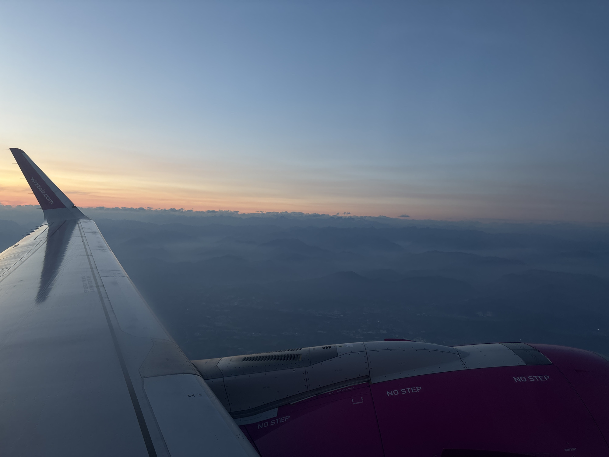 The image is an aerial view from an airplane window, likely captured during sunrise or sunset, given the soft, gradient sky with hues of blue, pink, and orange. In the foreground, a portion of the airplane's wing is visible, extending from the left edge of the frame. The wing is metallic and appears to be white or silver, reflecting the ambient light. A portion of the airplane's engine housing, painted in a vibrant magenta color, is visible in the lower right quadrant of the image. Text, including "NO STEP" and "wizzair.com," is visible on the wing and engine components.
Below the wing and engine, a landscape of rolling hills or mountains is visible, shrouded in a light mist or haze, creating a sense of depth and distance. Some faint signs of human habitation, such as scattered lights or structures, can be discerned within the darkened landscape, suggesting a rural or semi-urban area. The scene depicts a moment of flight over a serene, atmospheric landscape.
Regarding the location in the city of Caravaggio, Italy: While the image depicts a landscape that could be consistent with the geographical features of the Lombardy region where Caravaggio is located, there is no specific visual information within the image that definitively places it in Caravaggio itself. The landscape is generic enough to be many mountainous or hilly regions, and the aerial perspective does not offer identifiable landmarks of Caravaggio.